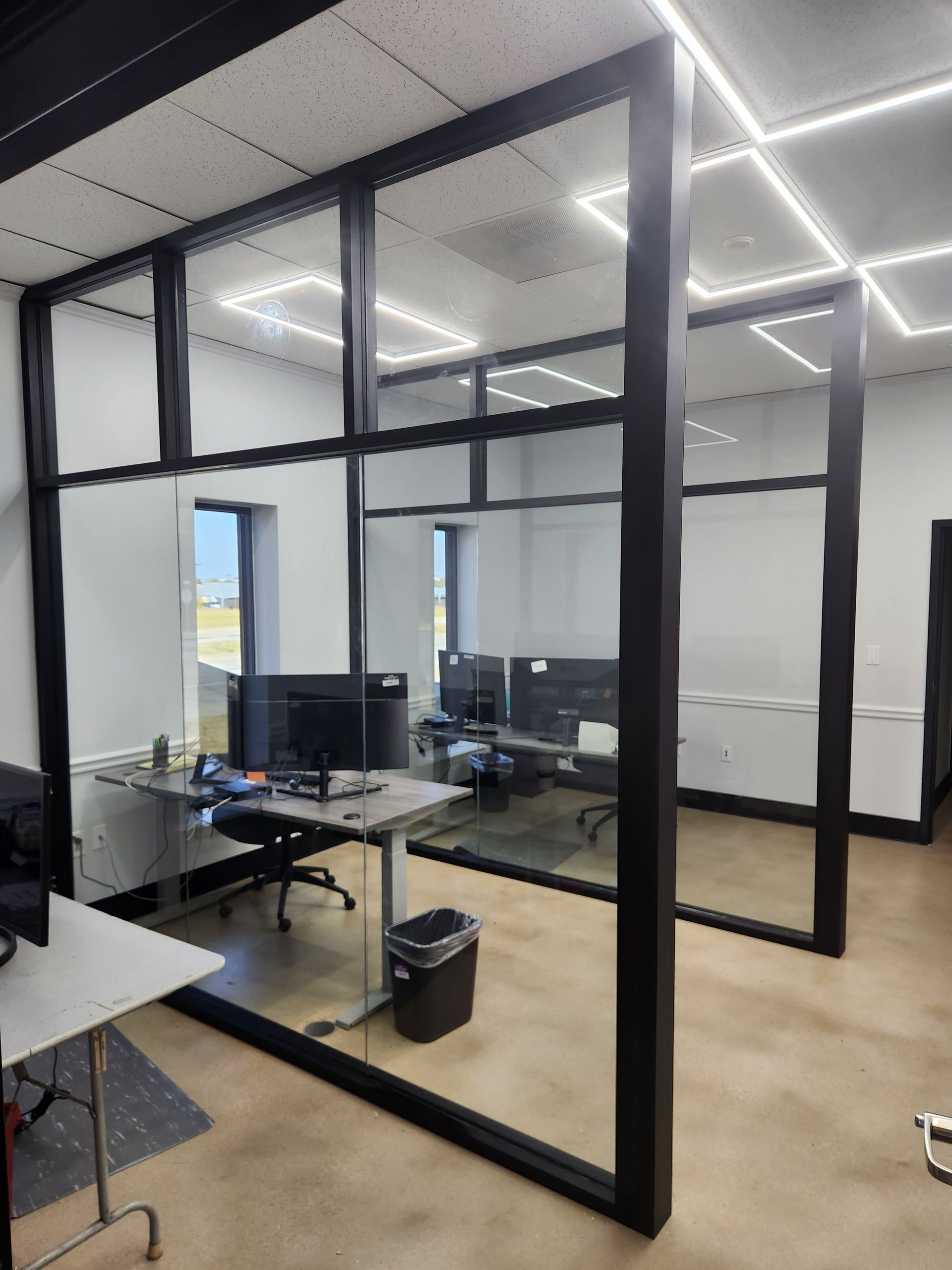 Office space with glass-walled cubicles. Black frames surround the glass. Desks with computers visible. Fluorescent lights on the ceiling.