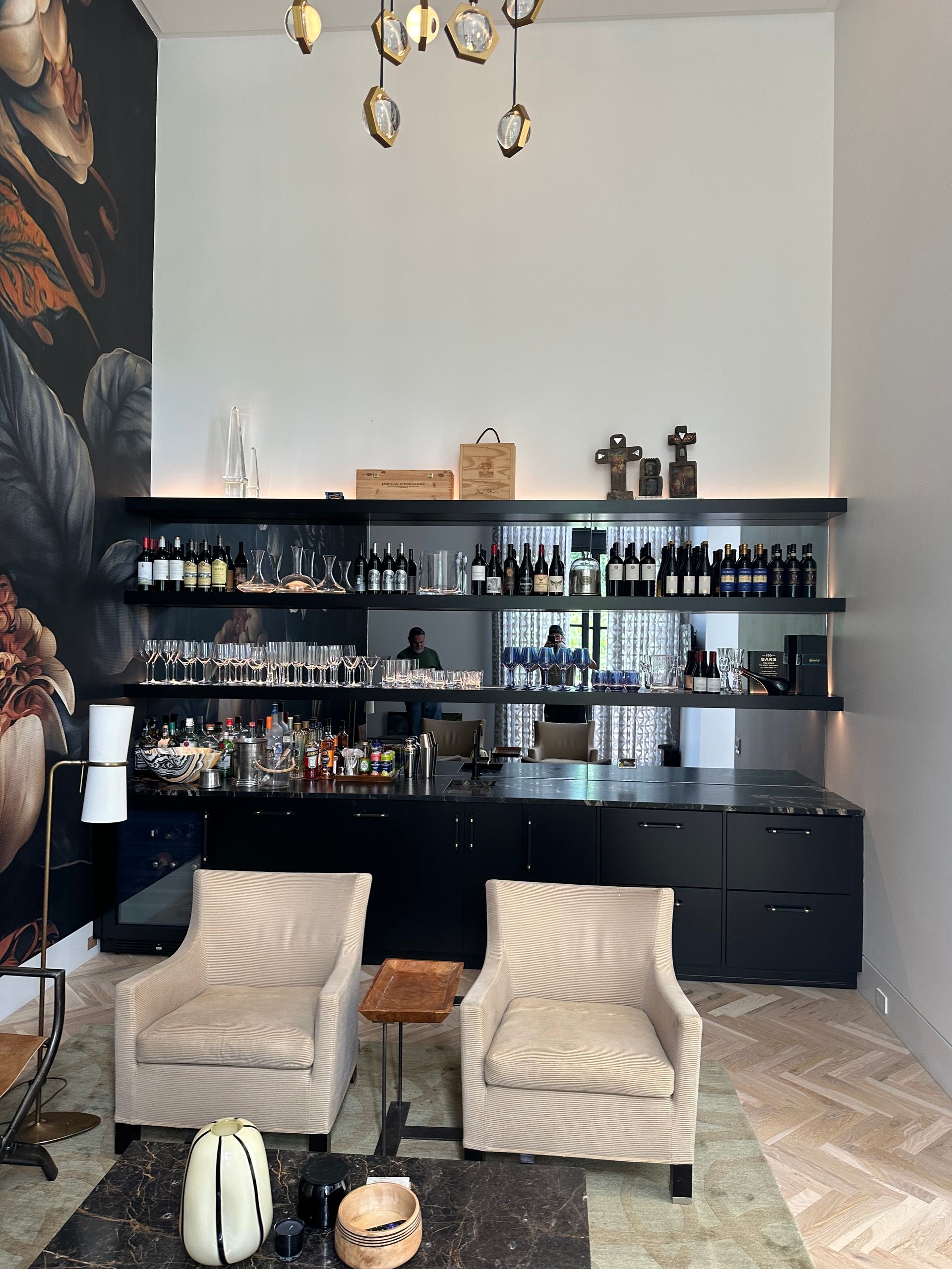 Lounge area with bar. Two cream upholstered chairs face a black bar with shelves of liquor and glassware.