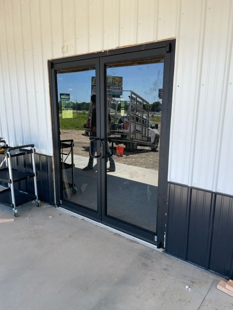 Two dark-framed glass doors reflecting a bright outdoor scene; the doors are set in a building with gray and white siding.