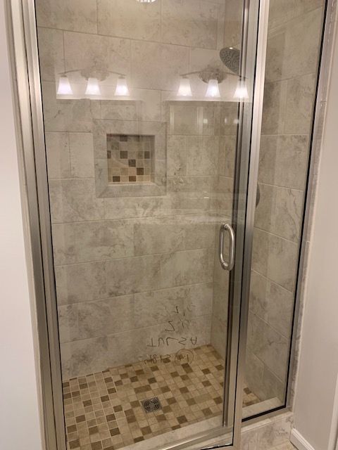 Shower with glass doors, silver frame, gray and brown tile, recessed shelf, overhead lighting, and handle.