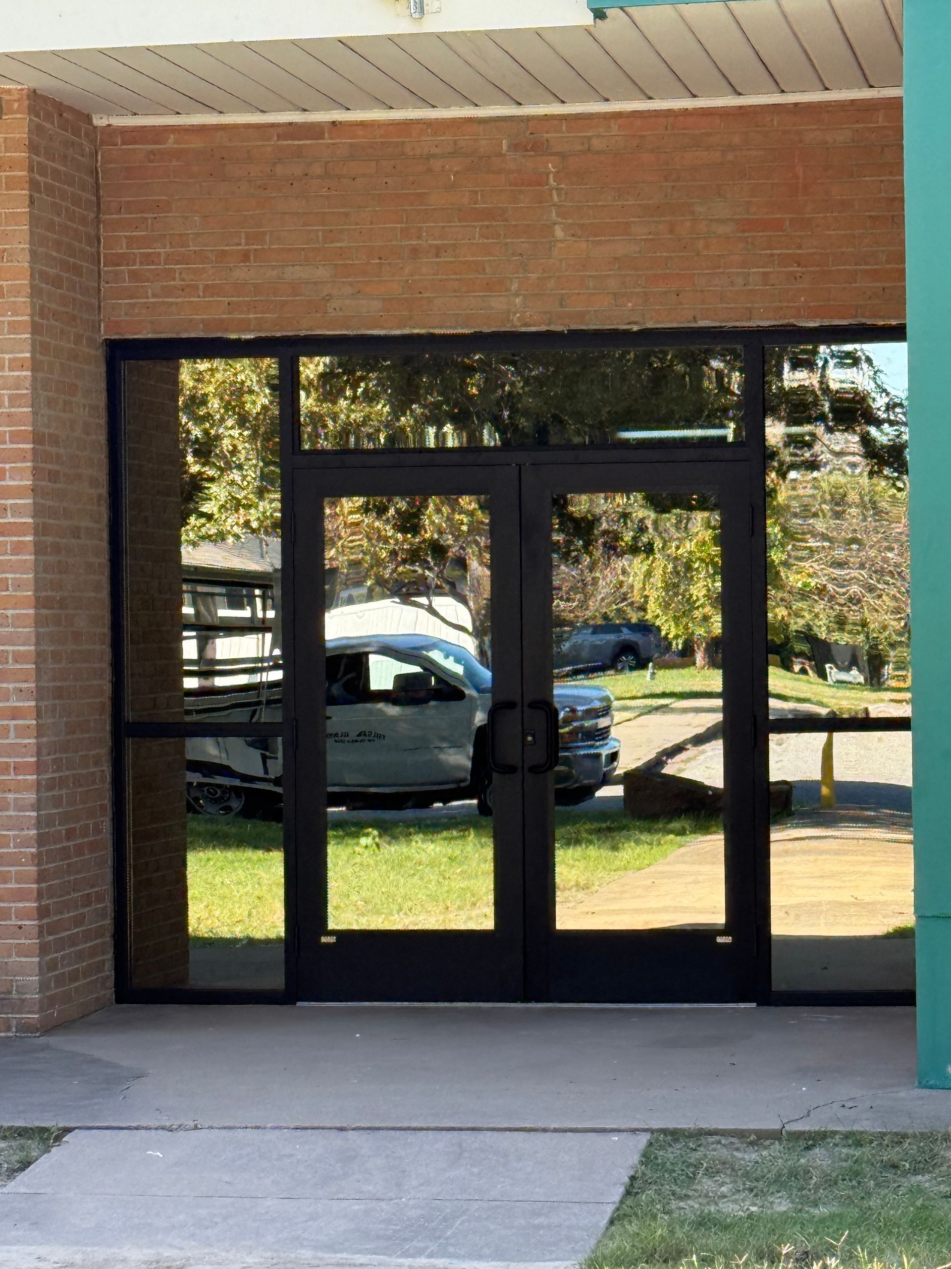 Glass double doors with black frames; brick building exterior. Reflection of a car and trees.