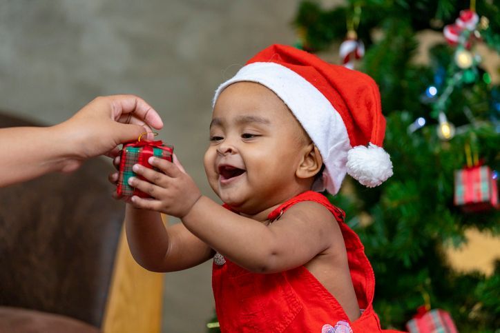 A baby wearing a santa hat is holding a gift box in front of a christmas tree.