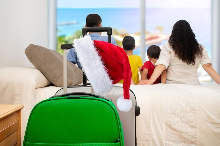A family is sitting on a bed with a green suitcase with a santa hat on it.