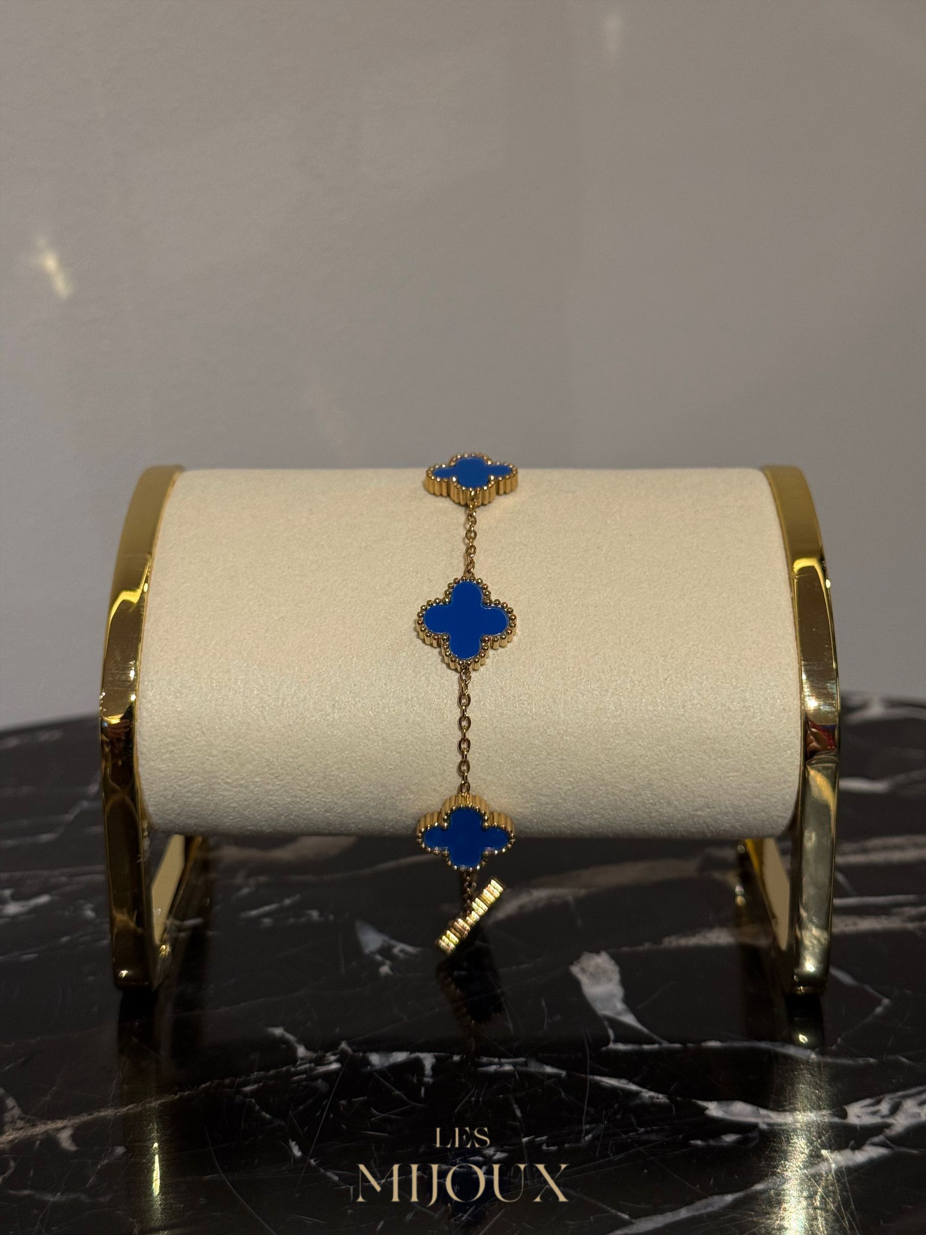 Gouden armband met drie blauwe klavermotieven, gepresenteerd op een crèmekleurige standaard met gouden rand.