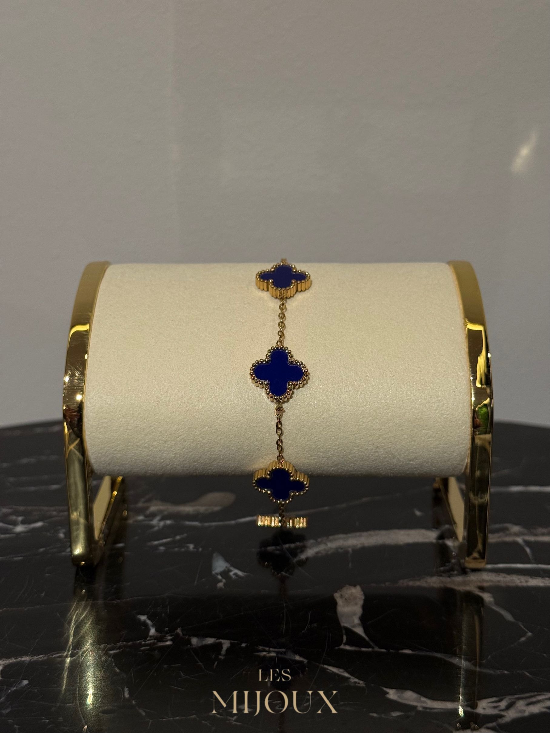 Gouden armband met blauwe klavervormige bedeltjes op een crèmekleurige ondergrond.
