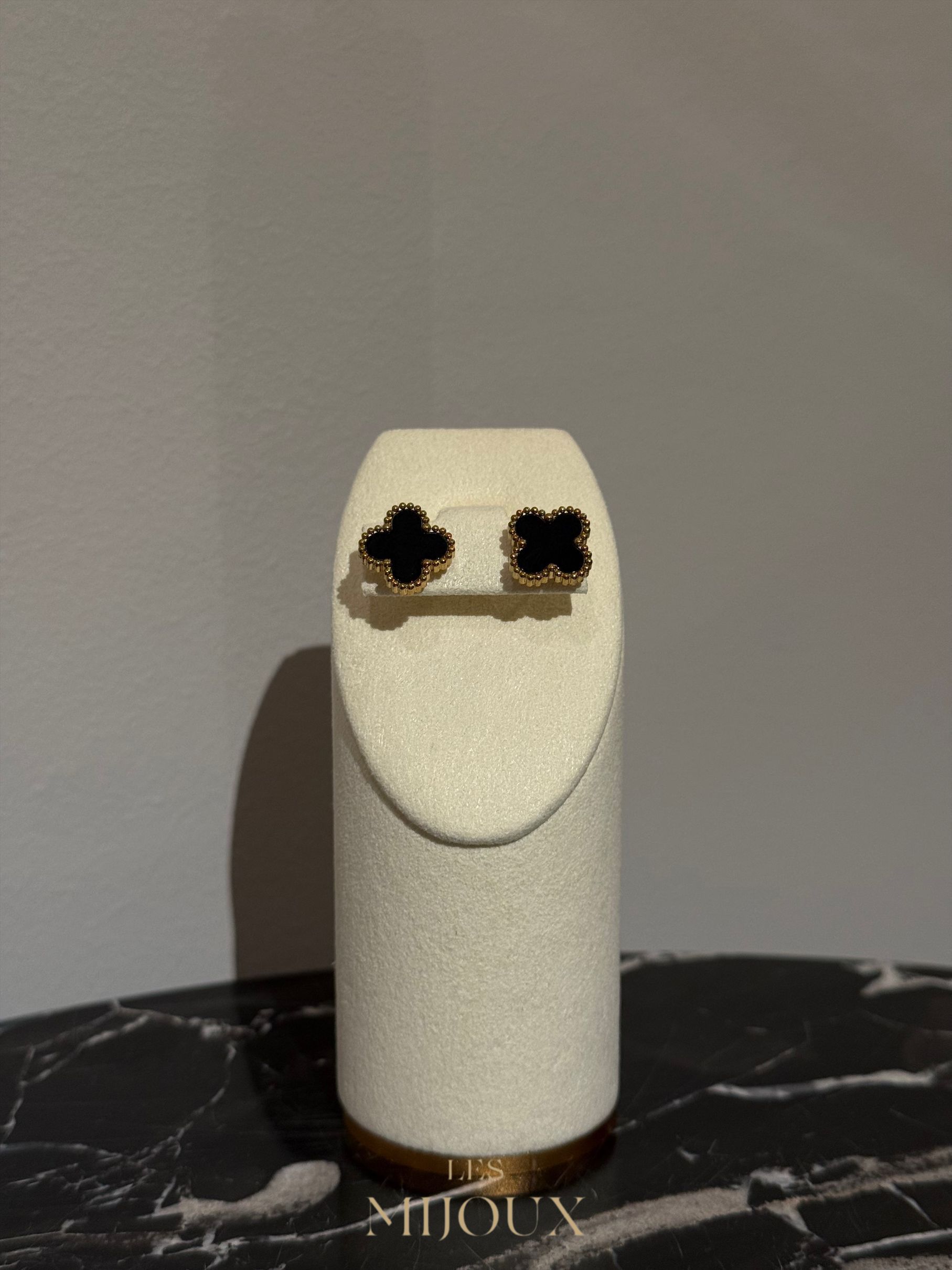 Zwarte klavervormige oorbellen op een witte sieradenstandaard tegen een grijze muur.