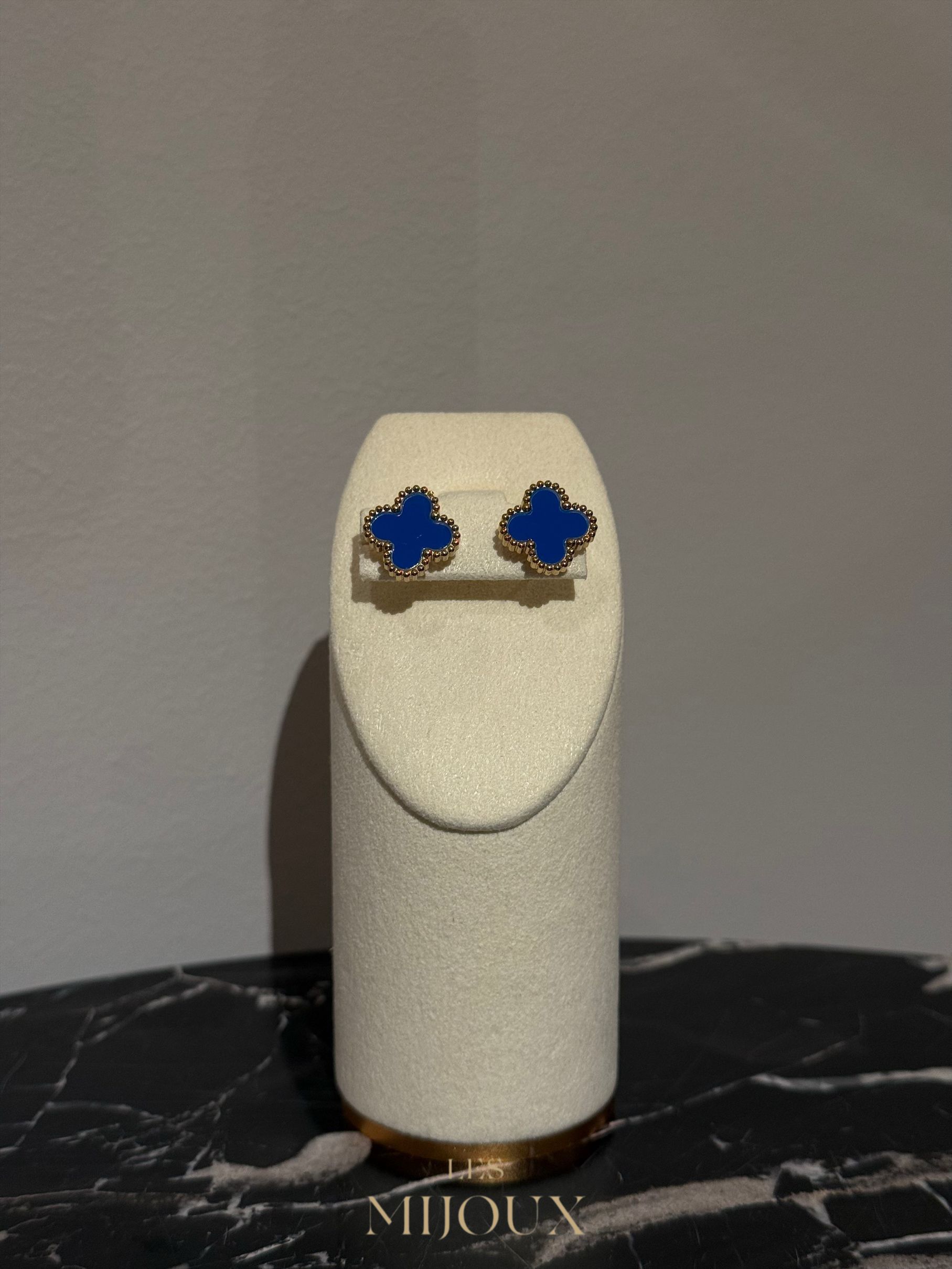 Blauwe klavervormige oorbellen op een witte sieradenstandaard, geplaatst tegen een grijze muur en marmeren ondergrond.