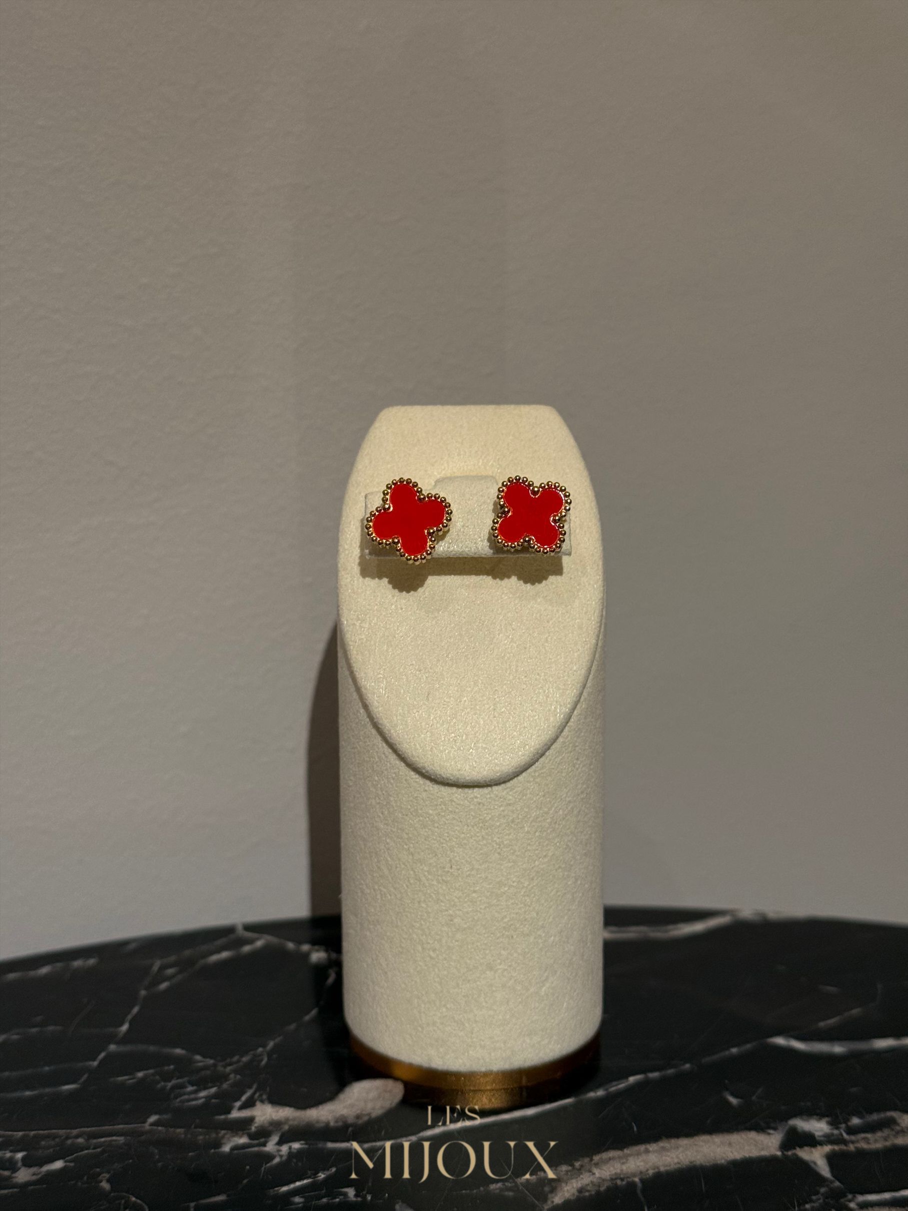 Oorbellen in de vorm van een rode klaver, op een crèmekleurige displaystandaard, geplaatst op een marmeren ondergrond.