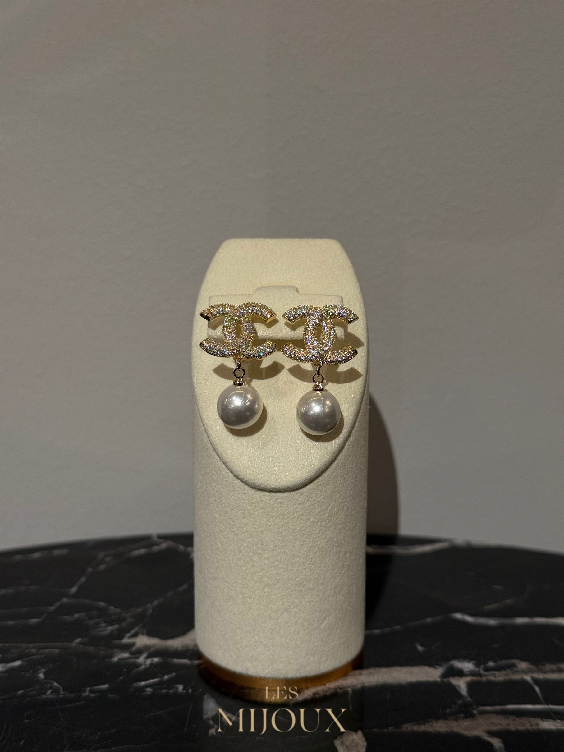 Chanel-oorbellen tentoongesteld op een witte sieradenstandaard met een parelhanger en het Chanel-logo.