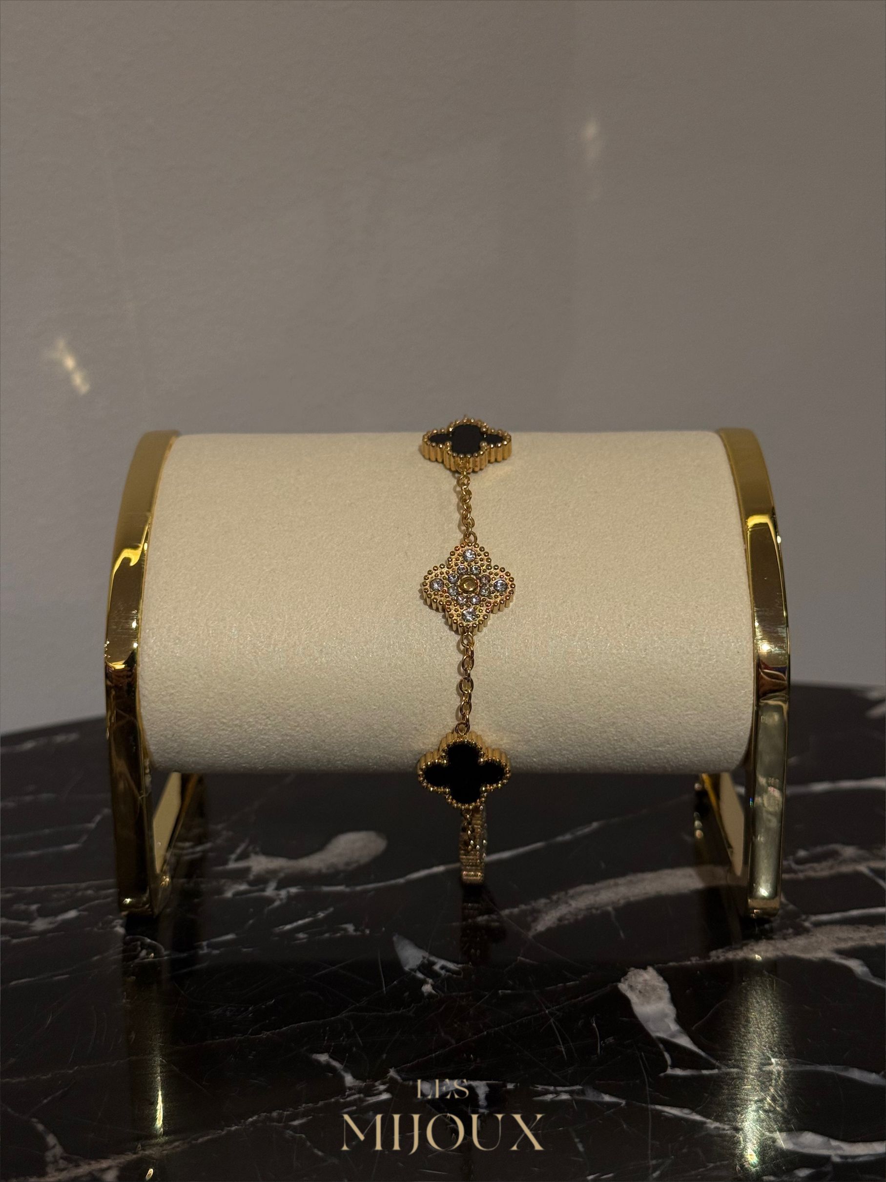 Gouden armband met zwarte klavervormige bedeltjes op een crèmekleurige standaard, geplaatst op een marmeren ondergrond.