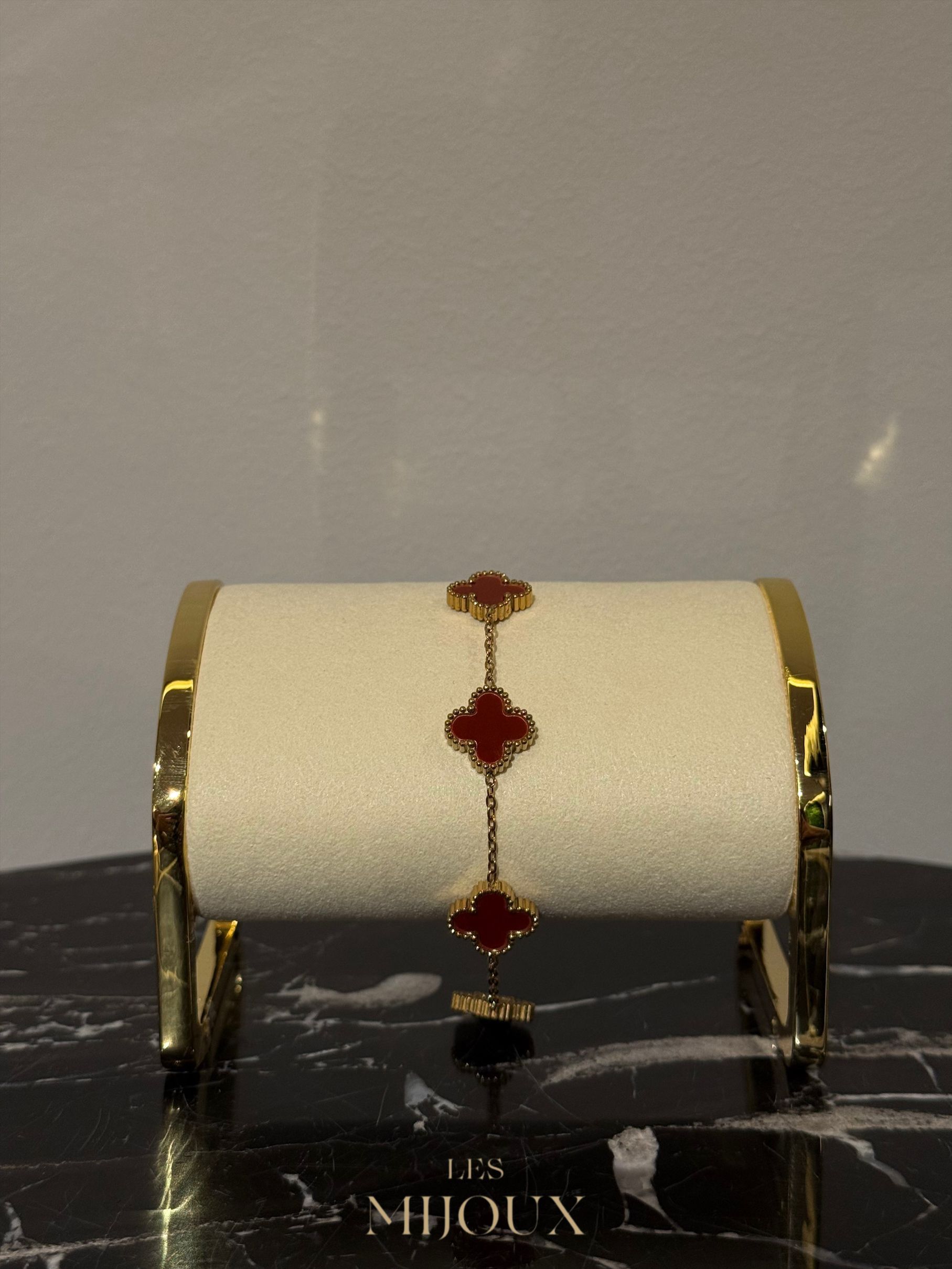 Gouden armband met drie rode klaverbedeltjes, gepresenteerd op een crèmekleurige standaard met een gouden rand.