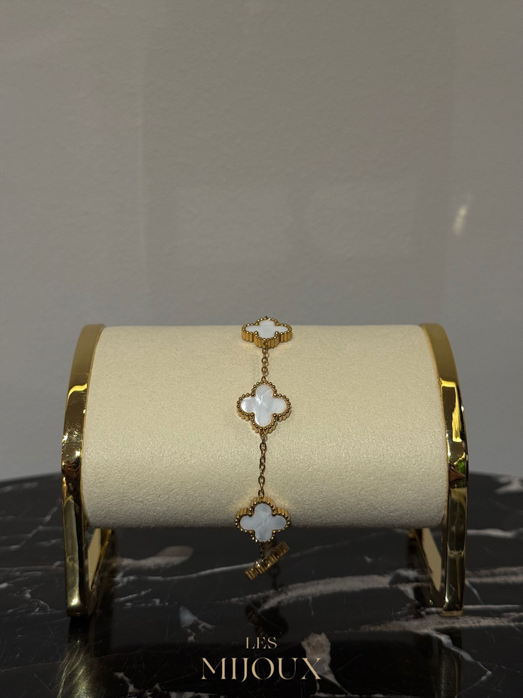 Gouden armband met drie klavervormige bedeltjes op een crèmekleurige displaystandaard.