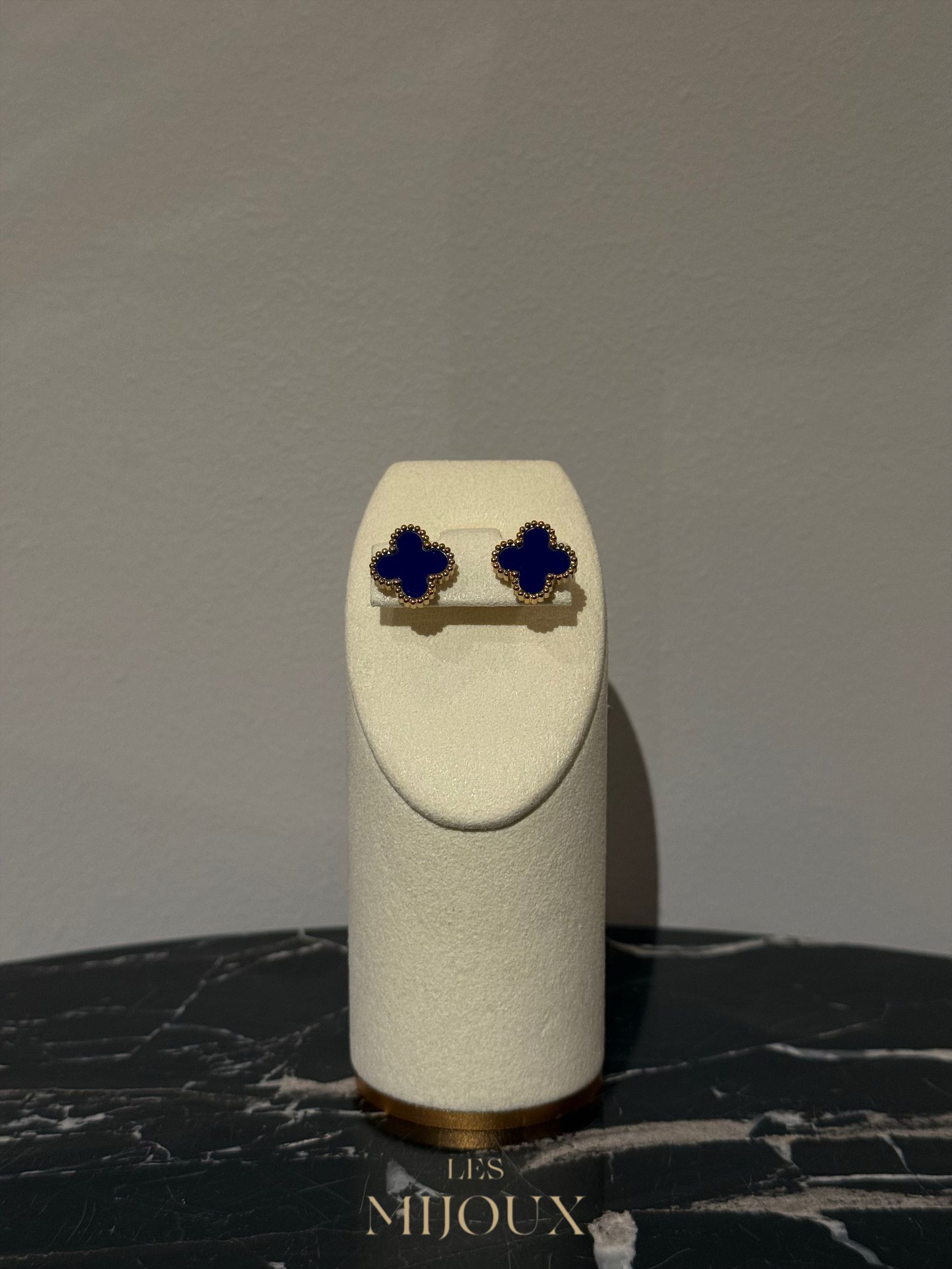 Blauwe klavervormige oorbellen op een crèmekleurige displaystandaard, geplaatst tegen een donkere marmeren achtergrond.