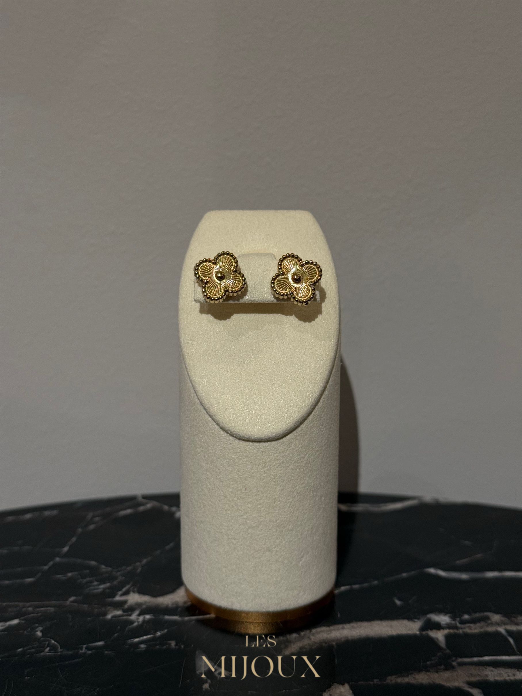 Gouden, bloemvormige oorknopjes op een witte sieradenstandaard tegen een gemarmerde achtergrond.