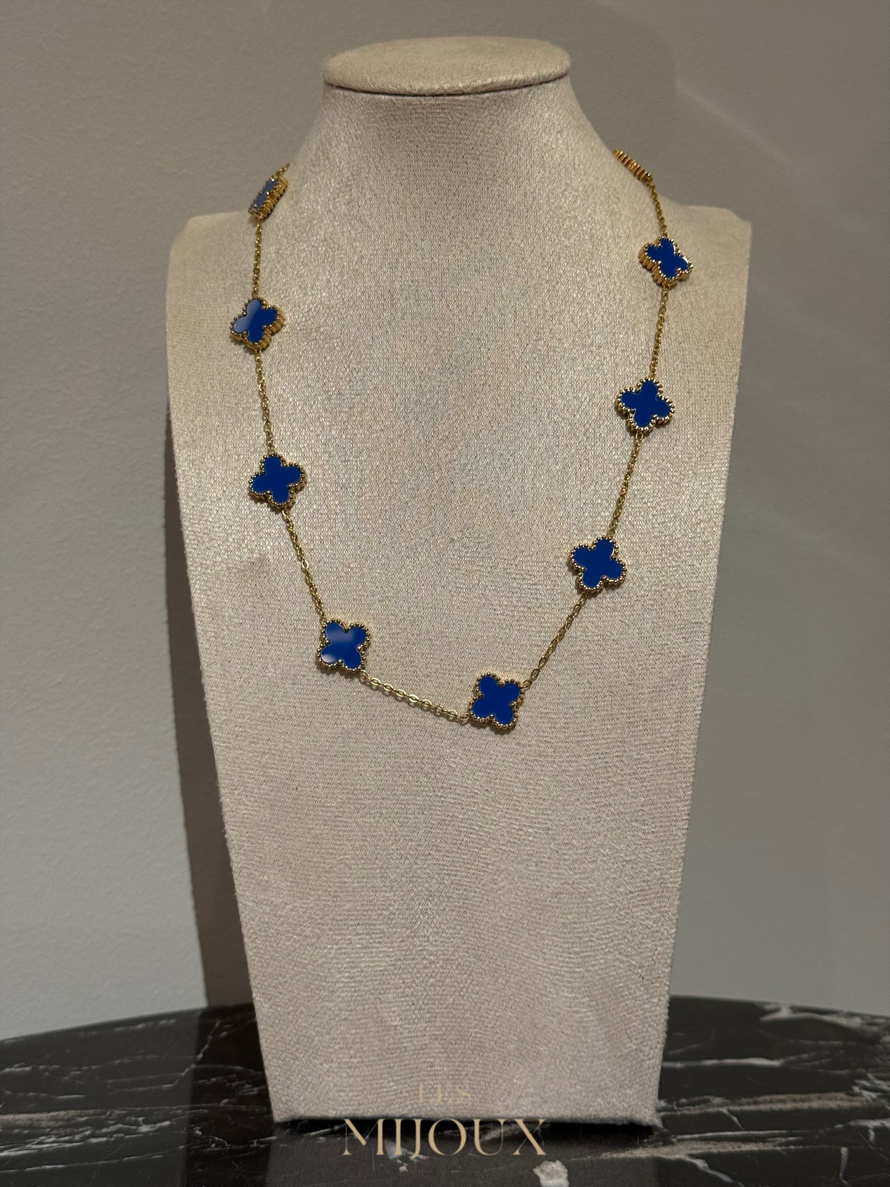 Gouden halsketting met blauwe klavervormige hangers, tentoongesteld op een beige sieradenbuste.