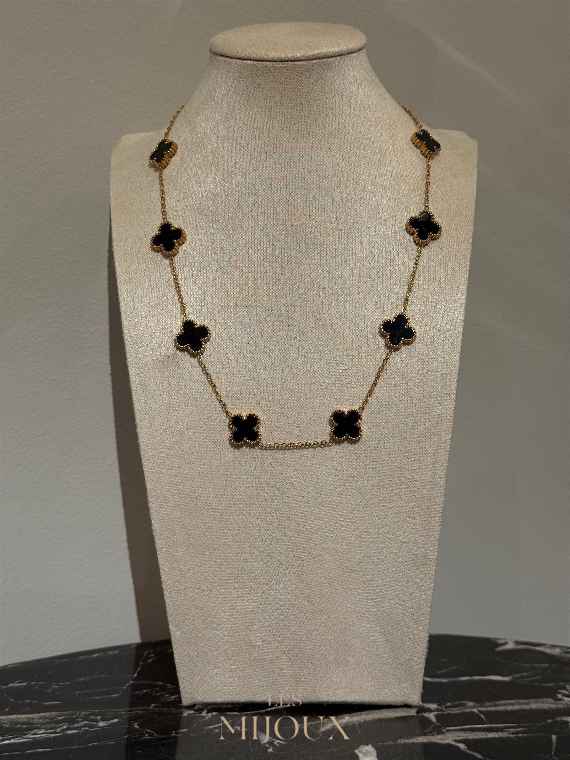 Gouden halsketting met zwarte klavervormige hangers op een beige displaystandaard.