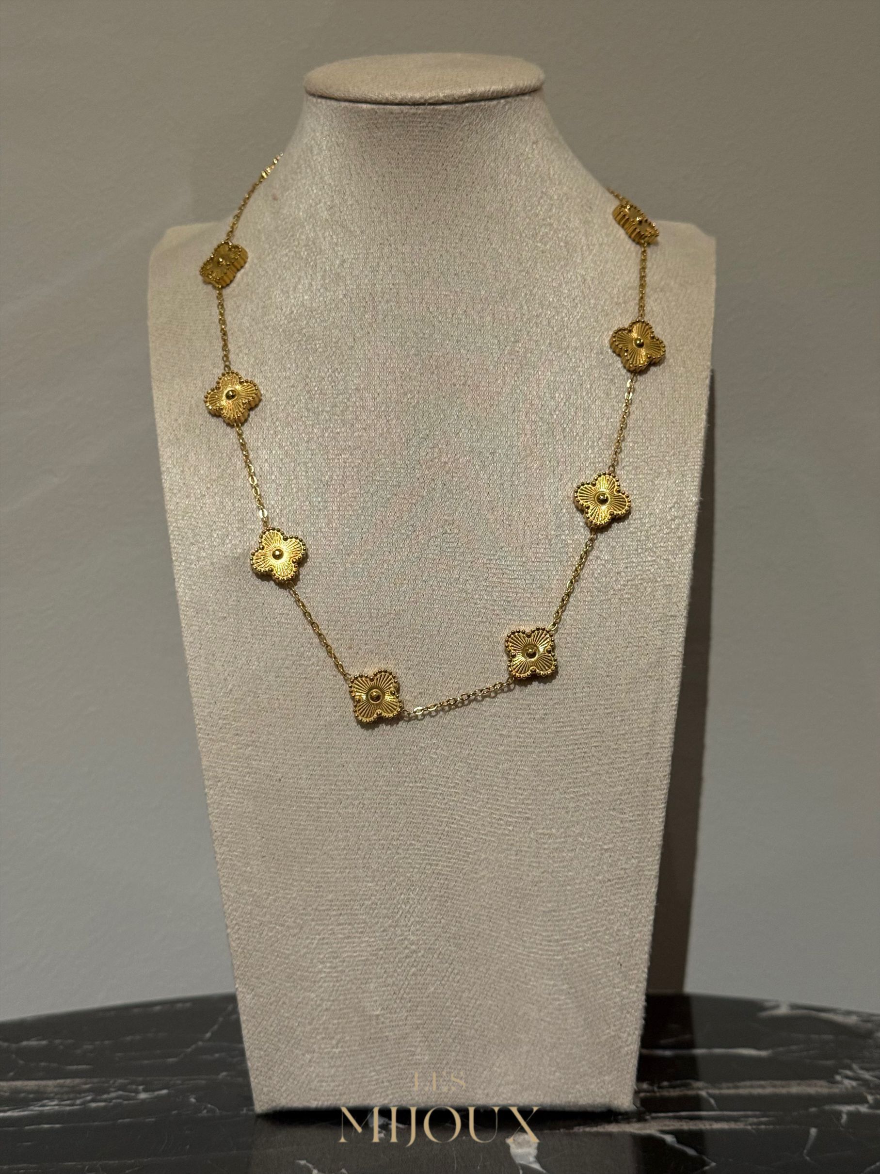 Gouden halsketting met bloemmotief, tentoongesteld op een crèmekleurige sieradenstandaard tegen een neutrale achtergrond.