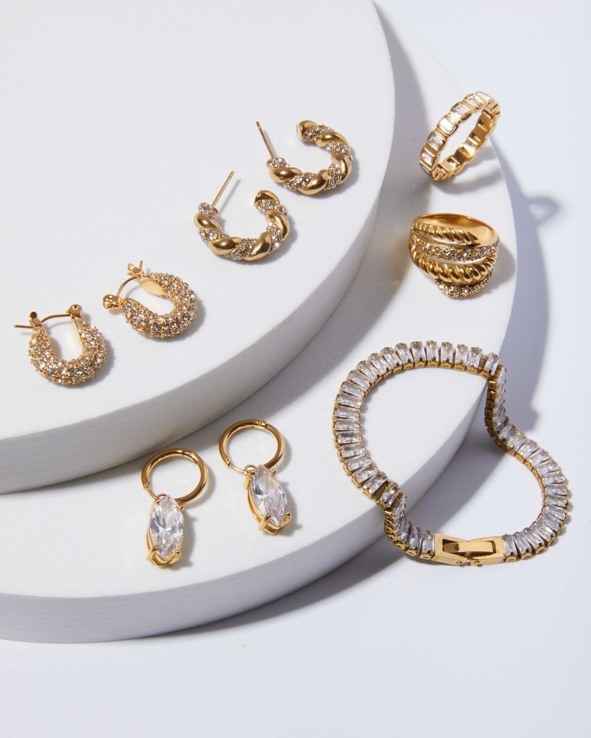 Gouden sieraden op een witte, trapsgewijs opgestelde ondergrond: oorbellen, ringen en een armband, die schitteren.