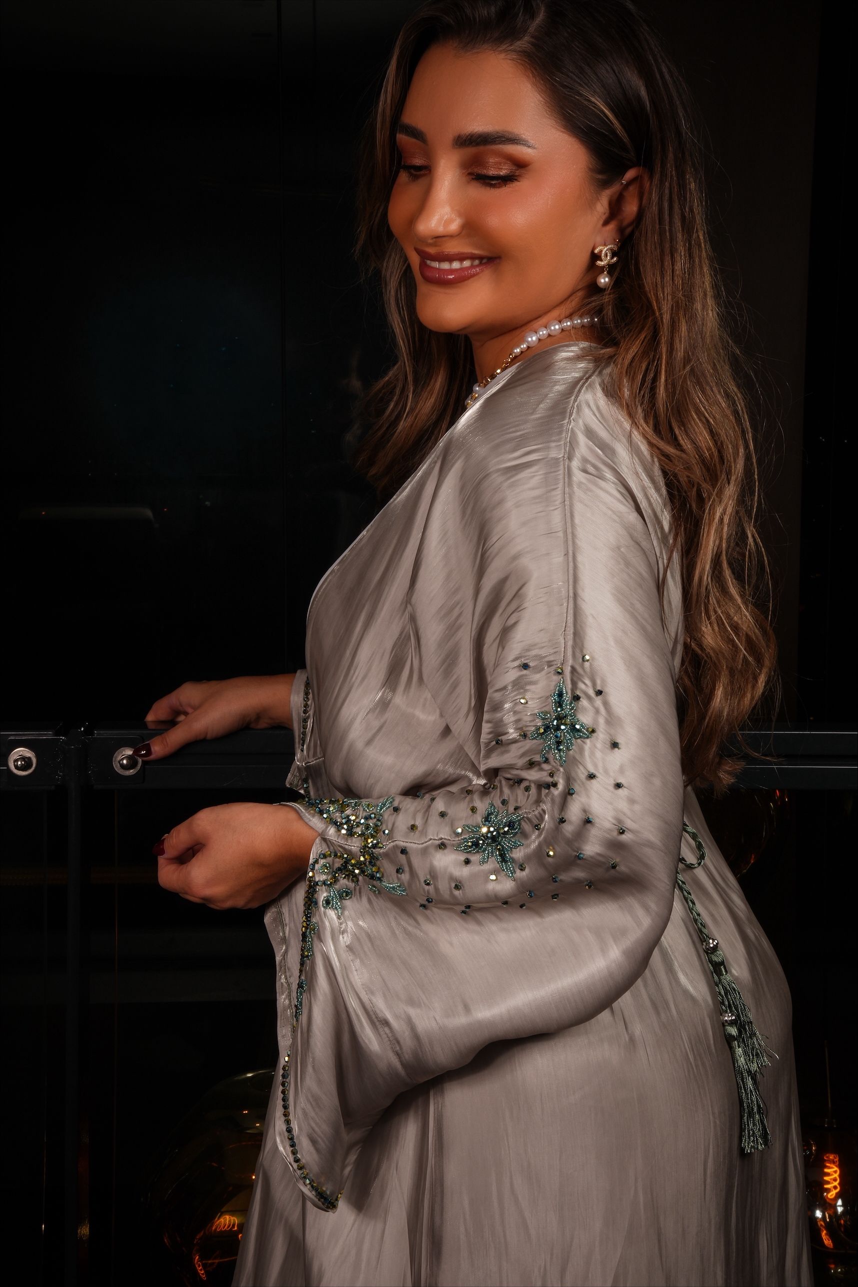 Vrouw in een grijze jurk met mouwen versierd met kralen, glimlachend, leunend tegen een reling in de nacht.