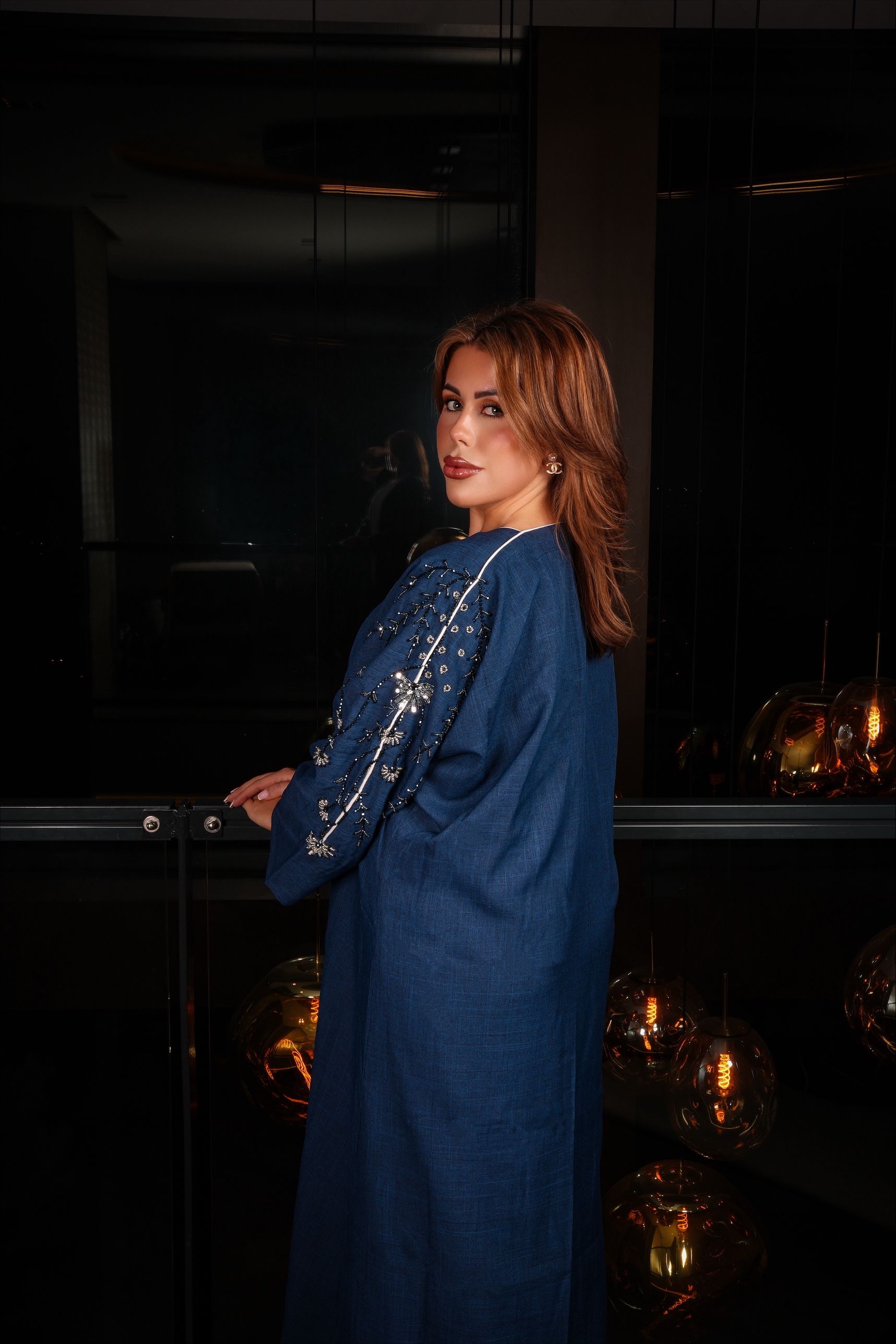 Vrouw in een lange, open blauwe jas over een witte jurk, staand bij een raam.