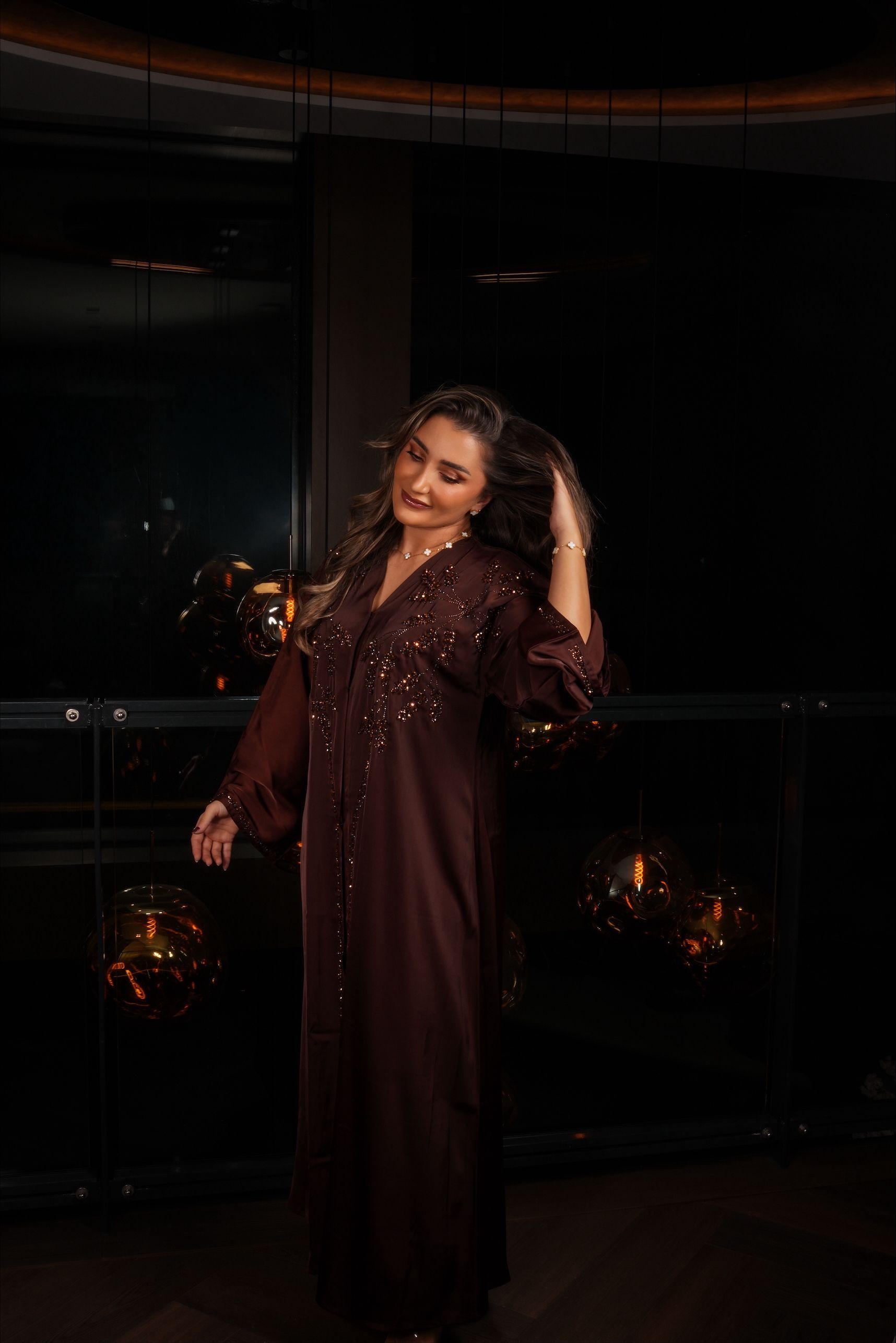 Vrouw in een bordeauxrode jurk, hand in haar haar, bij een raam met lampen.