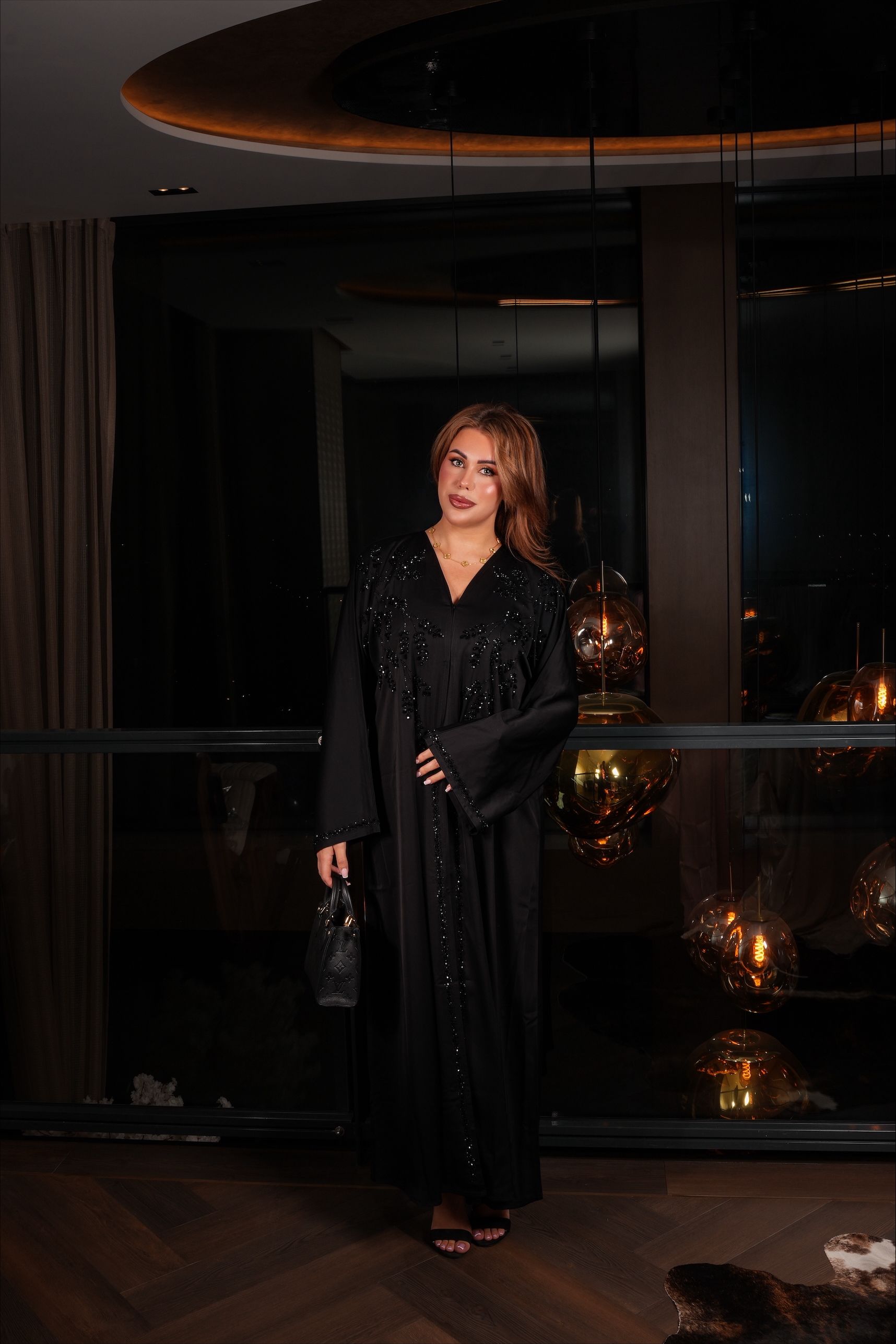 Vrouw in een zwarte, versierde jurk, met een handtas in haar hand, staat 's nachts bij een raam.