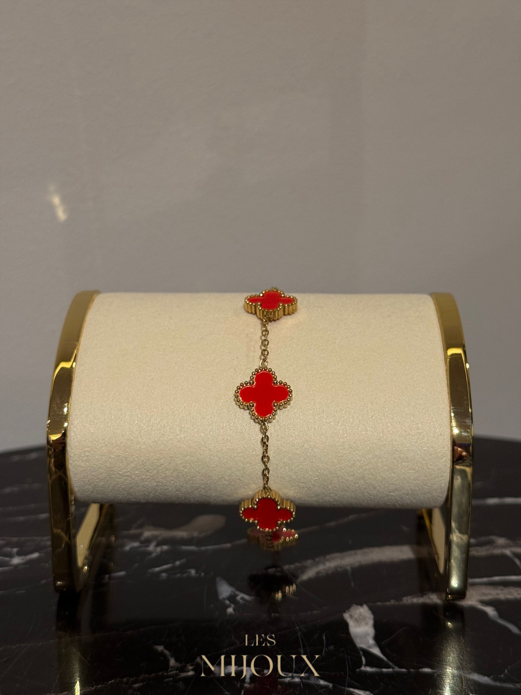 Gouden armband met drie rode klavervormige bedeltjes op een crèmekleurige cilinder.
