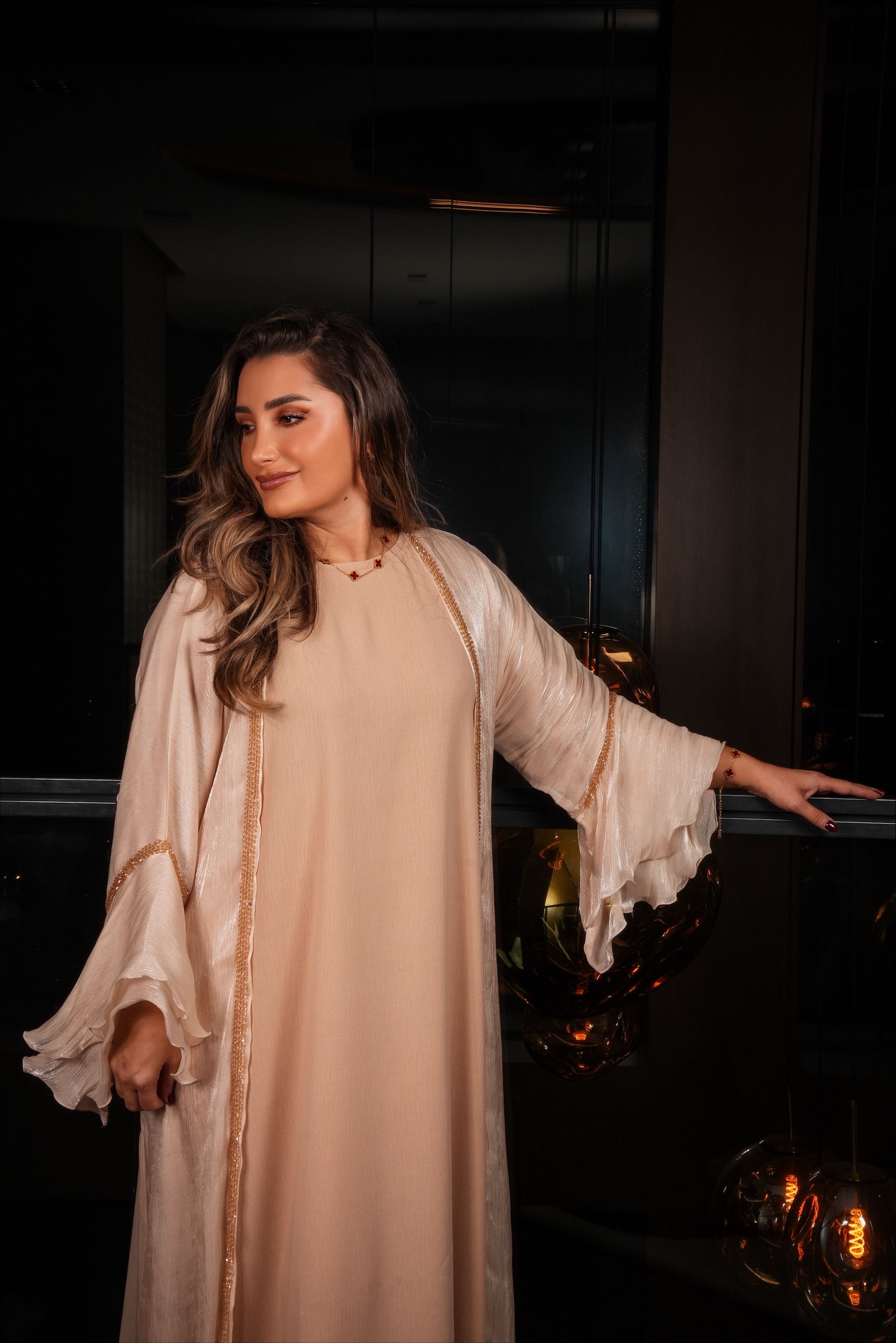 Vrouw in een beige jurk met pailletten, staand bij een donker raam.