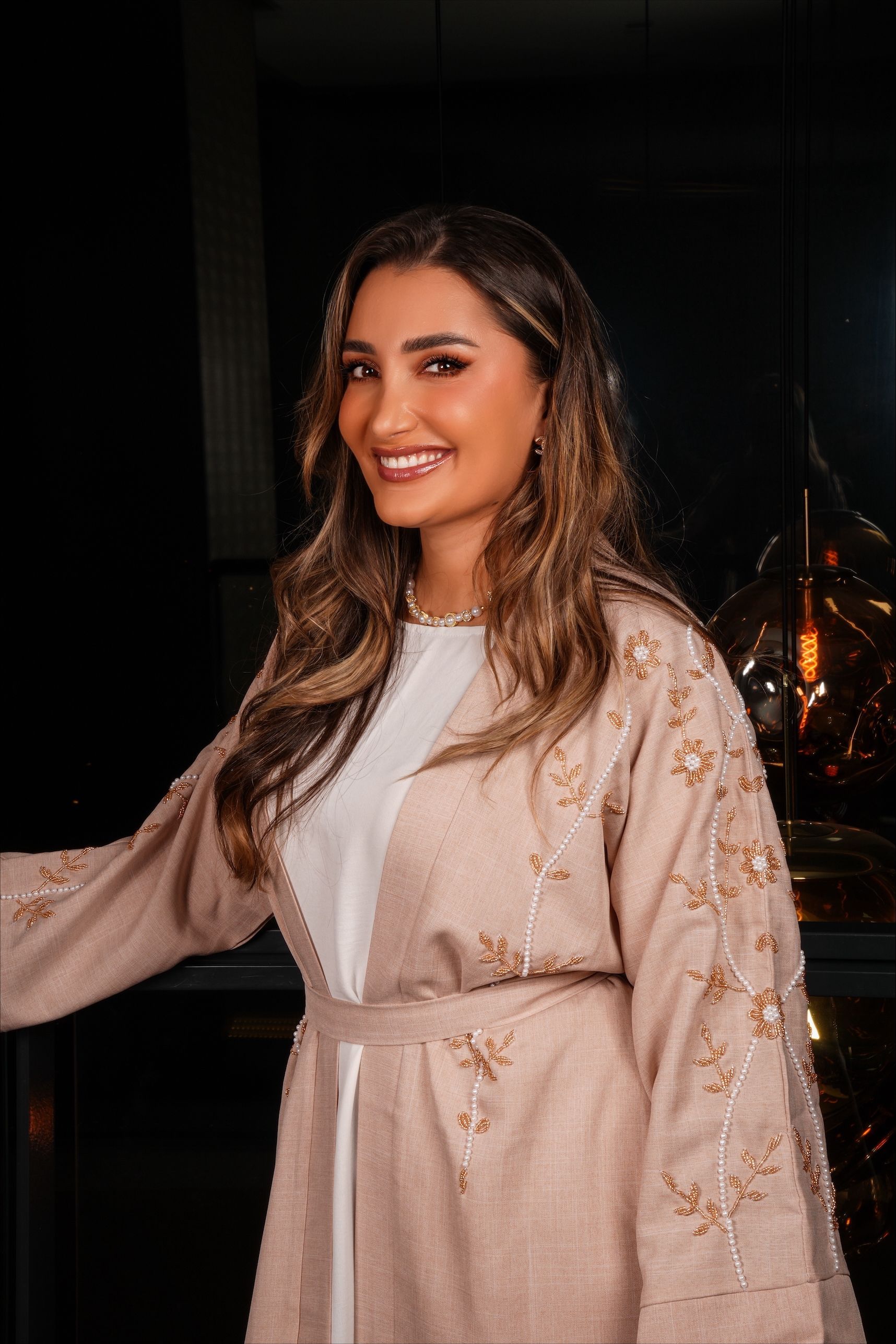 Vrouw in een beige jas met versieringen, leunend tegen een glazen balustrade met een donkere achtergrond.