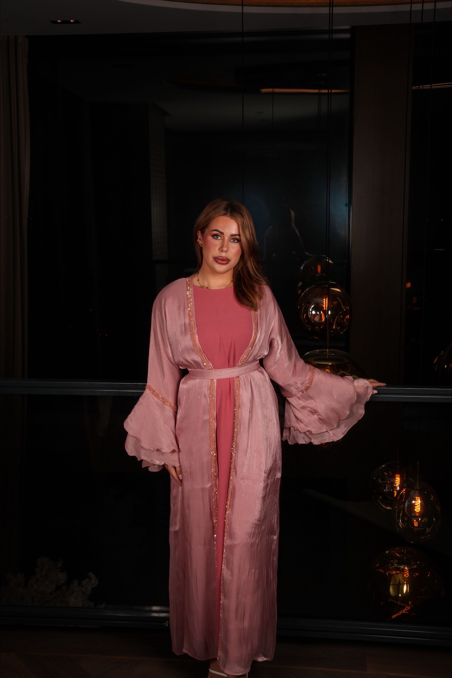 Een vrouw in een roze jurk en ochtendjas staat bij een donker raam.