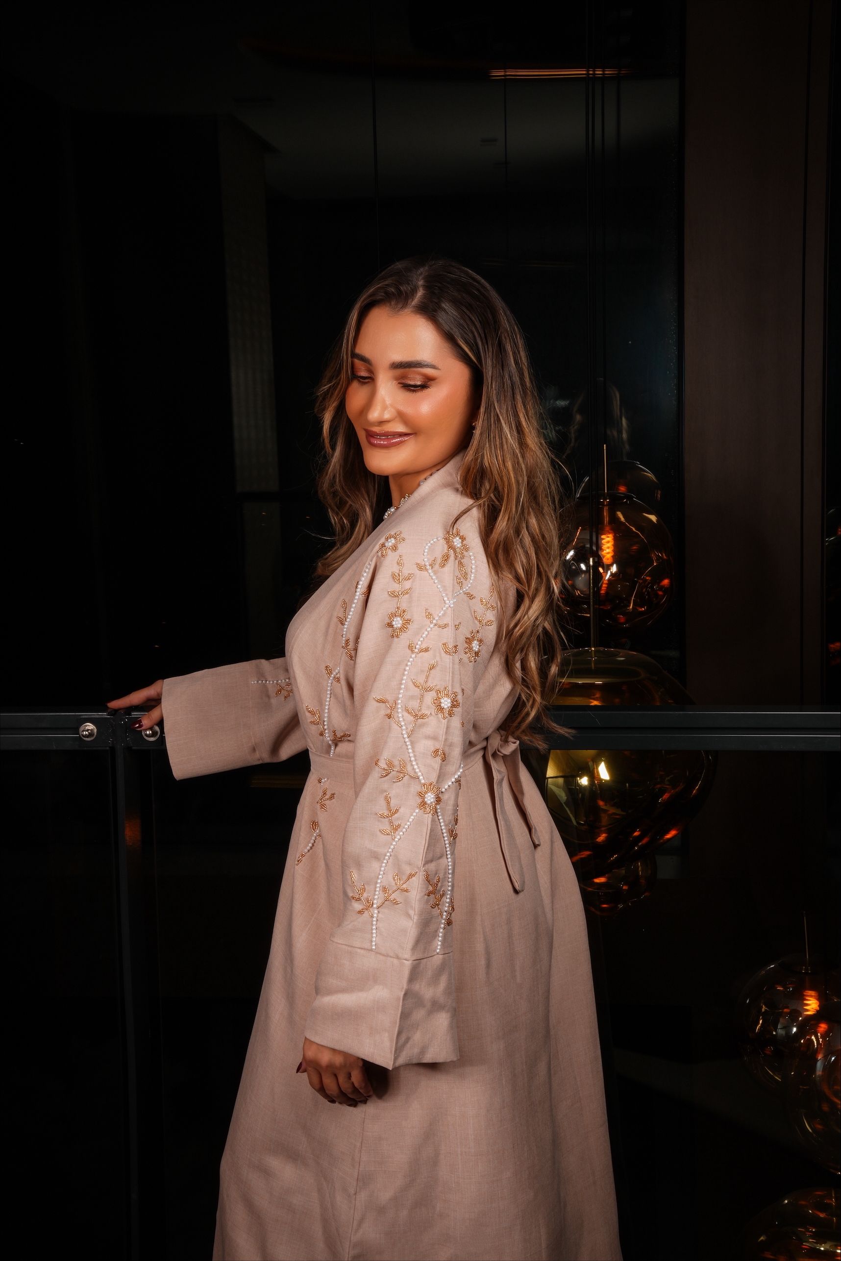 Vrouw in een beige jas met versieringen, leunend tegen een glazen balustrade met een donkere achtergrond.