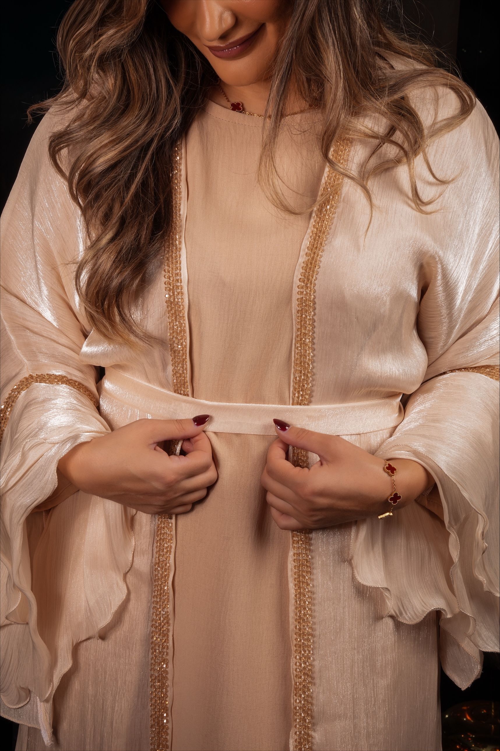 Vrouw in een beige jurk met pailletten, staand bij een donker raam.