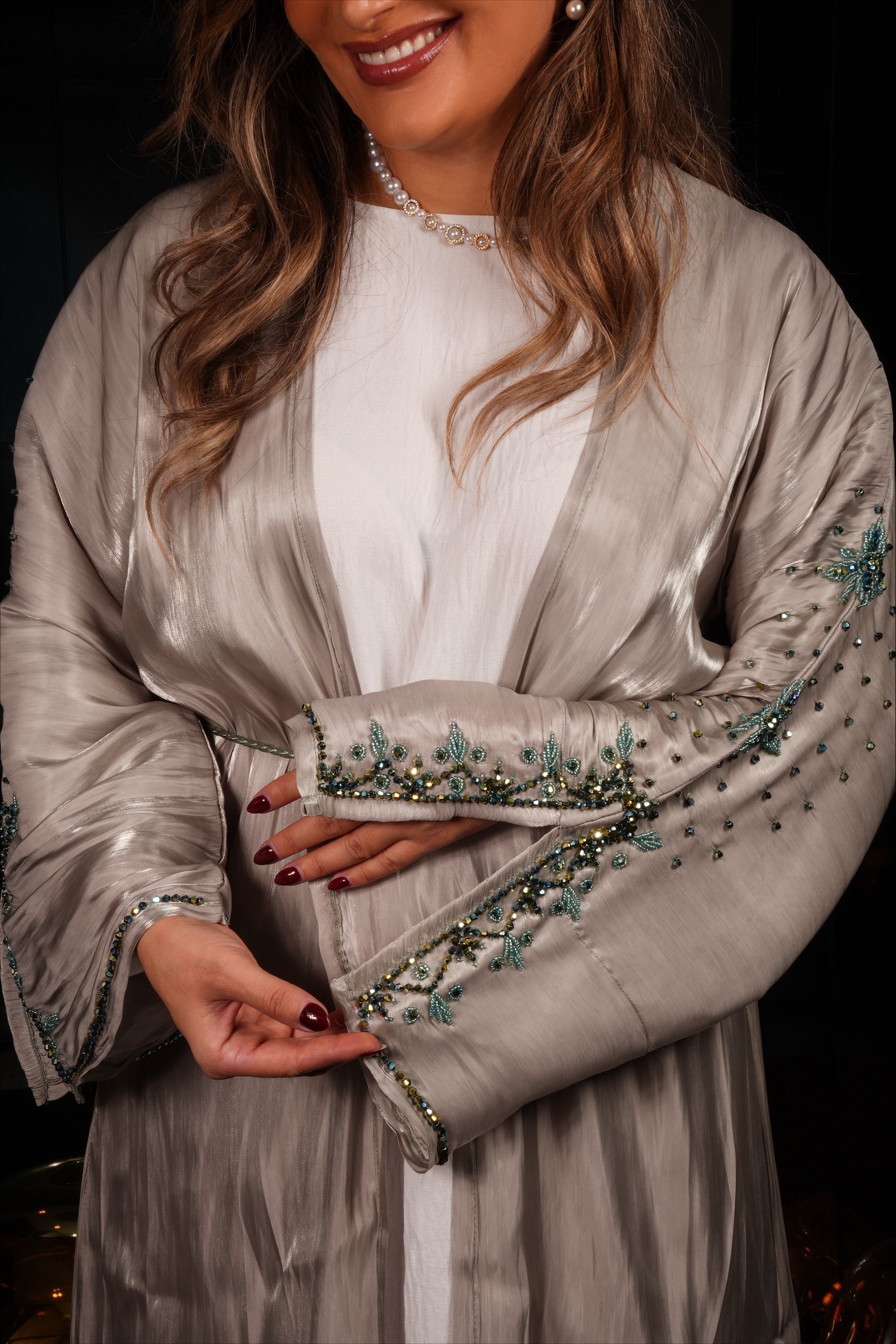 Vrouw in een grijze jurk met mouwen versierd met kralen, glimlachend, leunend tegen een reling in de nacht.