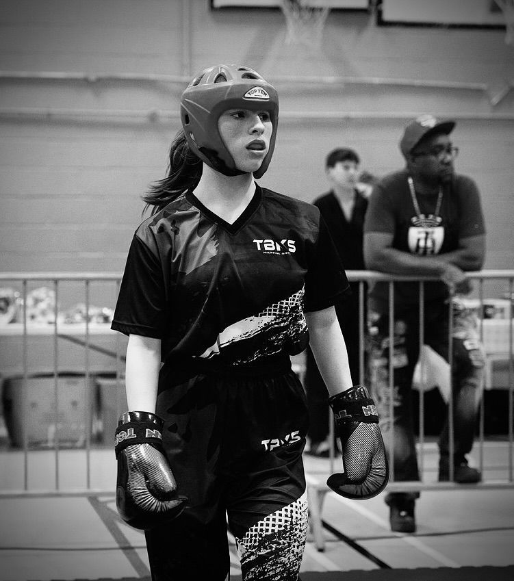 A young female martial artist in gear, helmet, and gloves, walks toward the viewer at a competition.