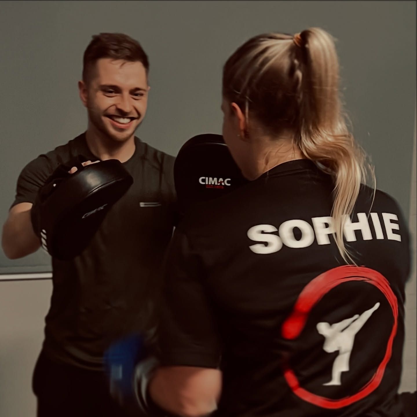 Man smiles, holding pads for a woman in a black shirt labeled