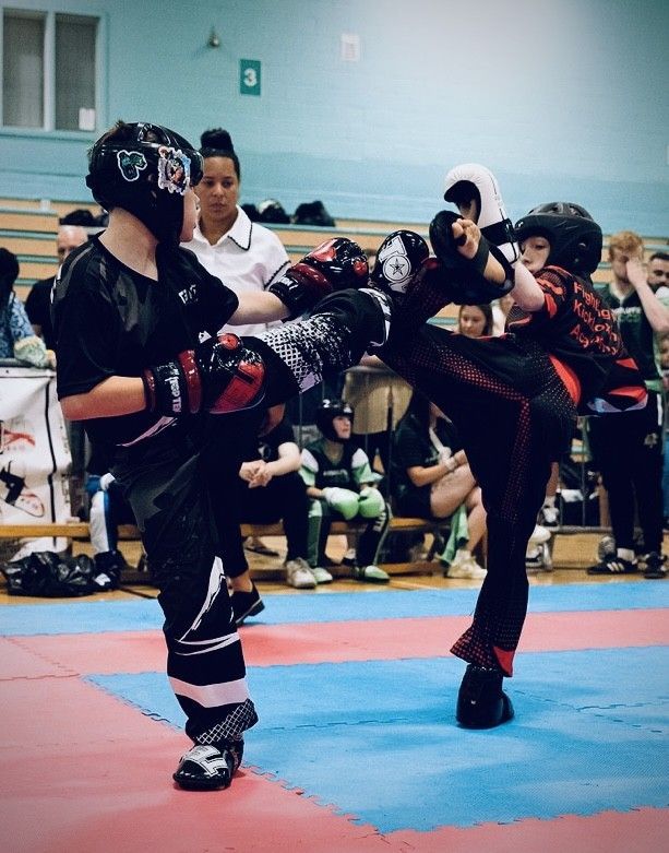 Two young kickboxers in black and red gear kick in a ring, watched by an official and spectators.