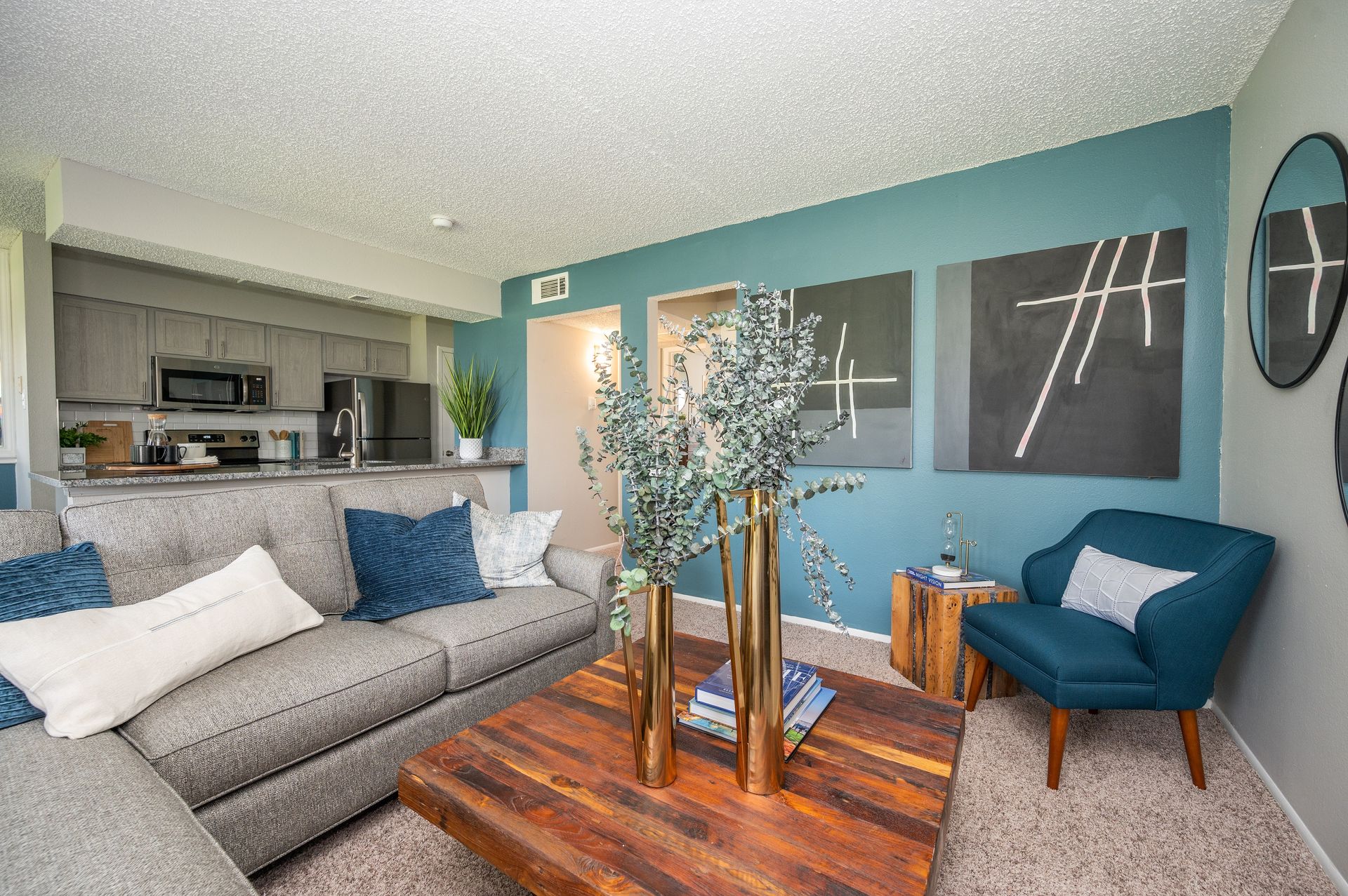 A modern living room with a gray sofa, teal accent wall, and a wooden coffee table in front of a kitchenette.