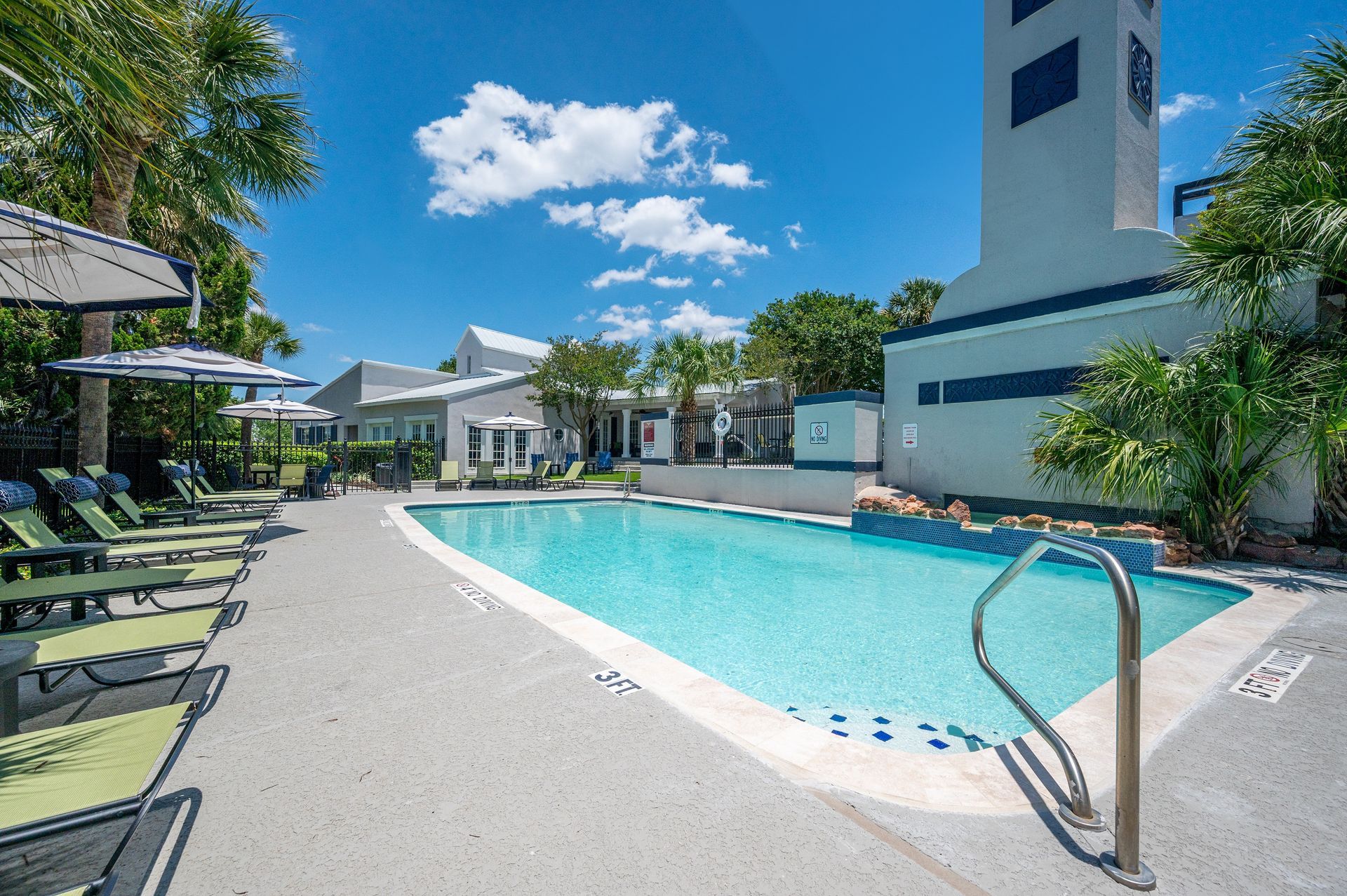 Pool with lounge chairs and umbrellas on a sunny day. A tall tower is visible near the pool's edge.