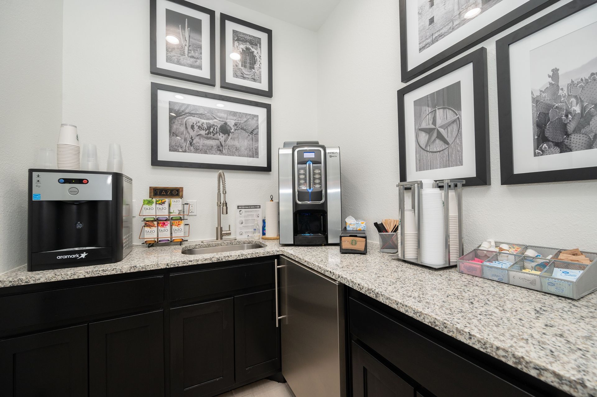 A small office kitchen area with a water cooler, coffee maker, and sink. Black cabinets and white walls are accented with black and white framed art.