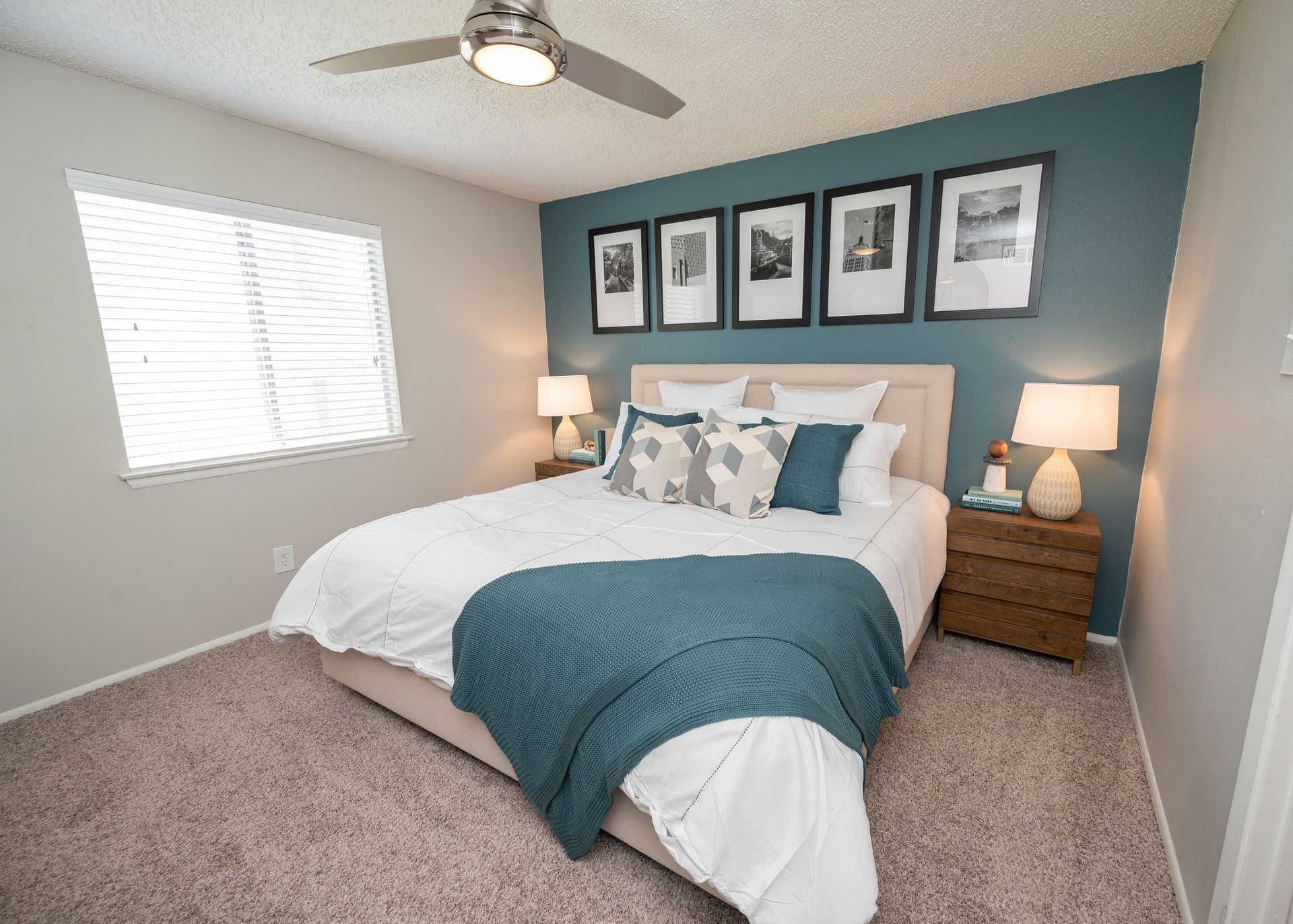 Bedroom with a teal accent wall, bed, nightstands, and framed photos. The room has a neutral carpet and blinds on the window.