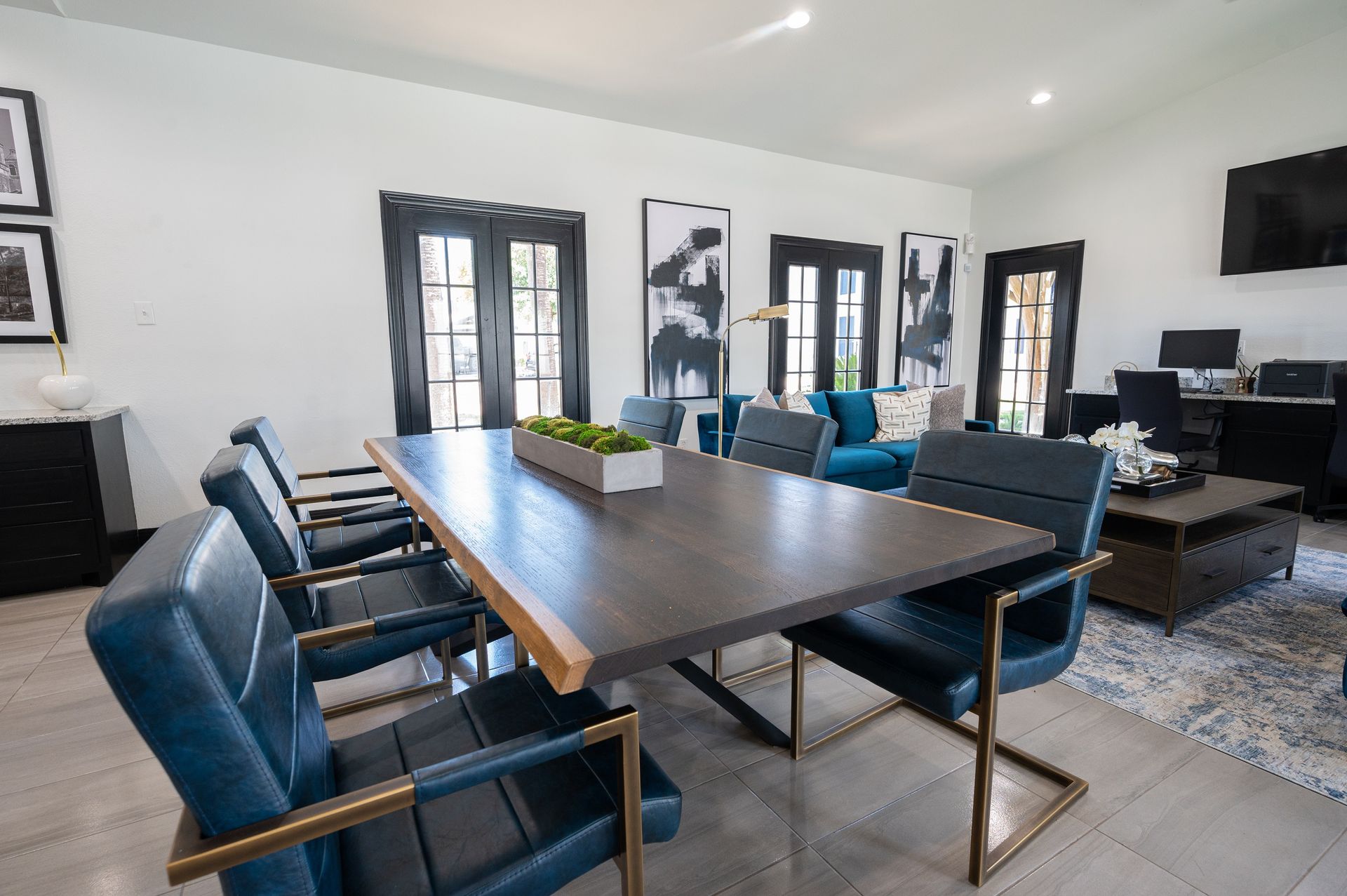 A dining room with a large wooden table and blue chairs, with black framed doors and artwork on the white walls.