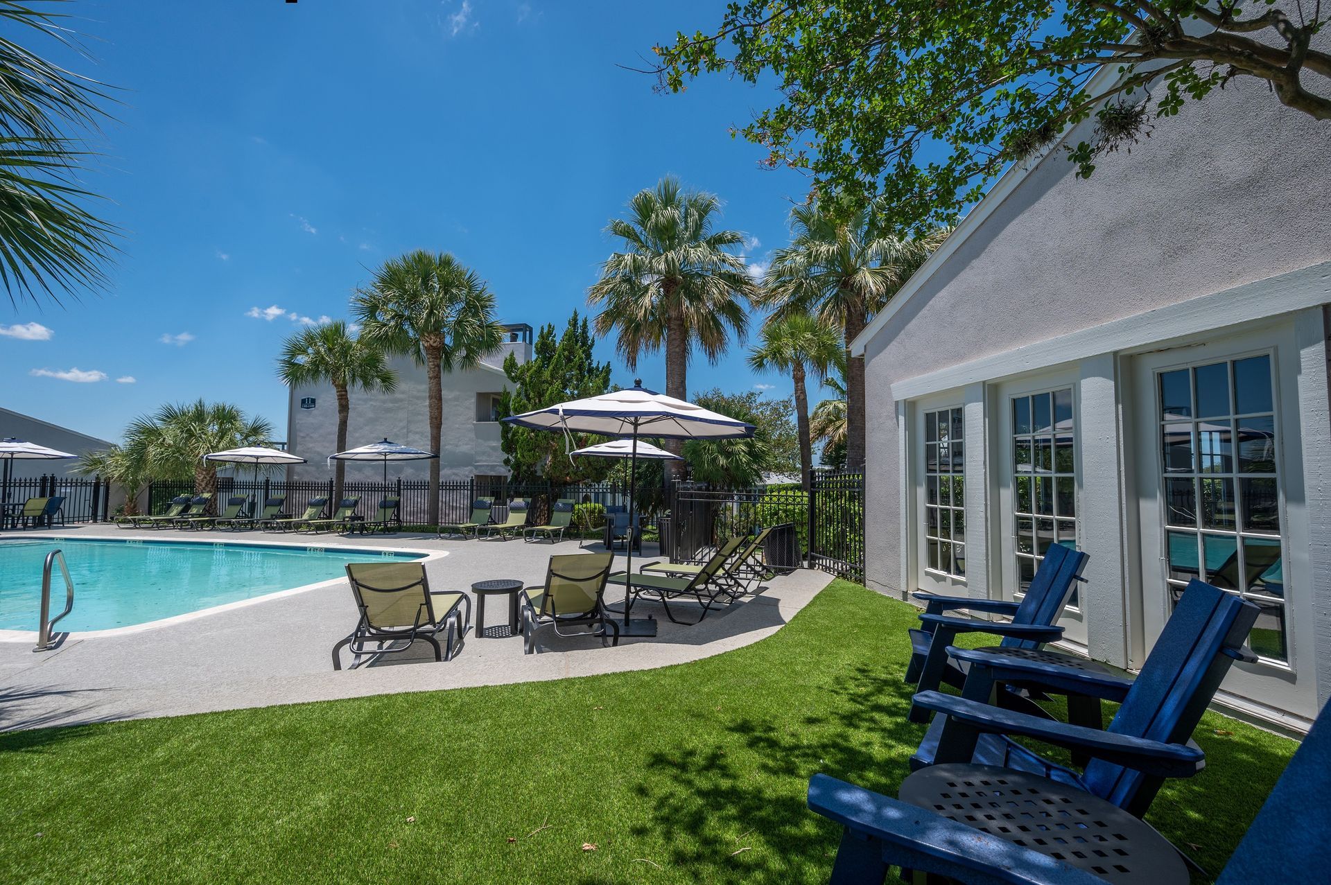 A pool area with lounge chairs, umbrellas, and a building with windows. Green grass and palm trees under a blue sky.