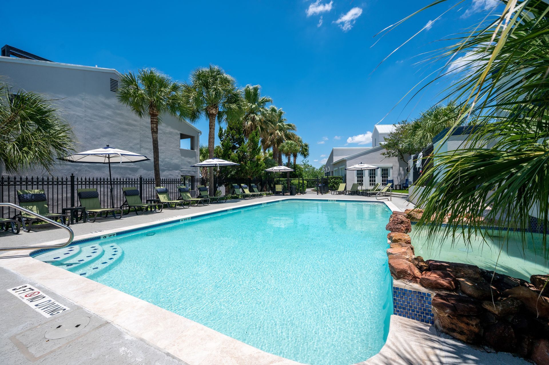 A swimming pool surrounded by palm trees and buildings under a bright blue sky. The water is a vibrant turquoise.