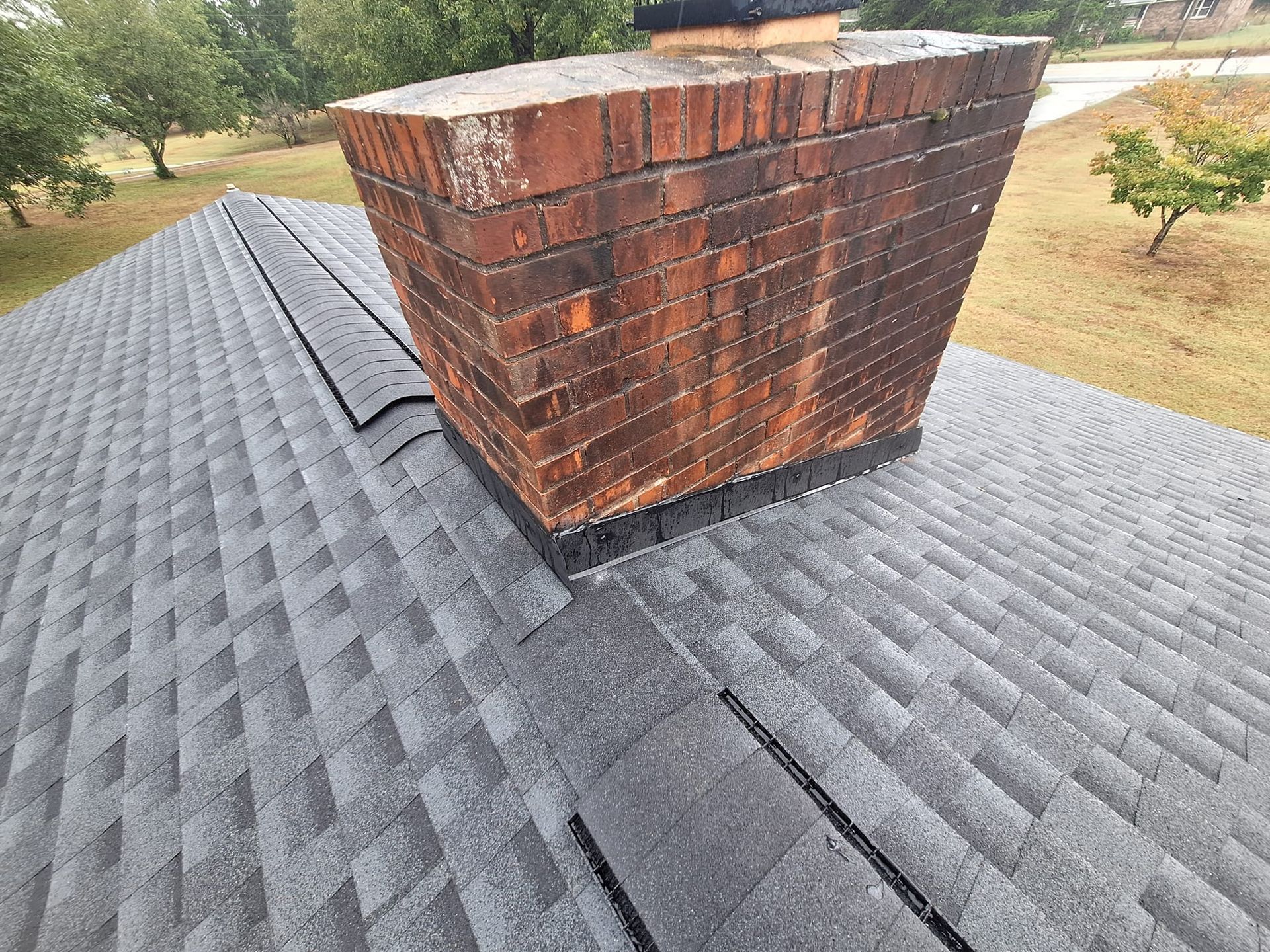 Brick chimney on a gray shingle roof, showing water stains. Overcast day in yard setting.