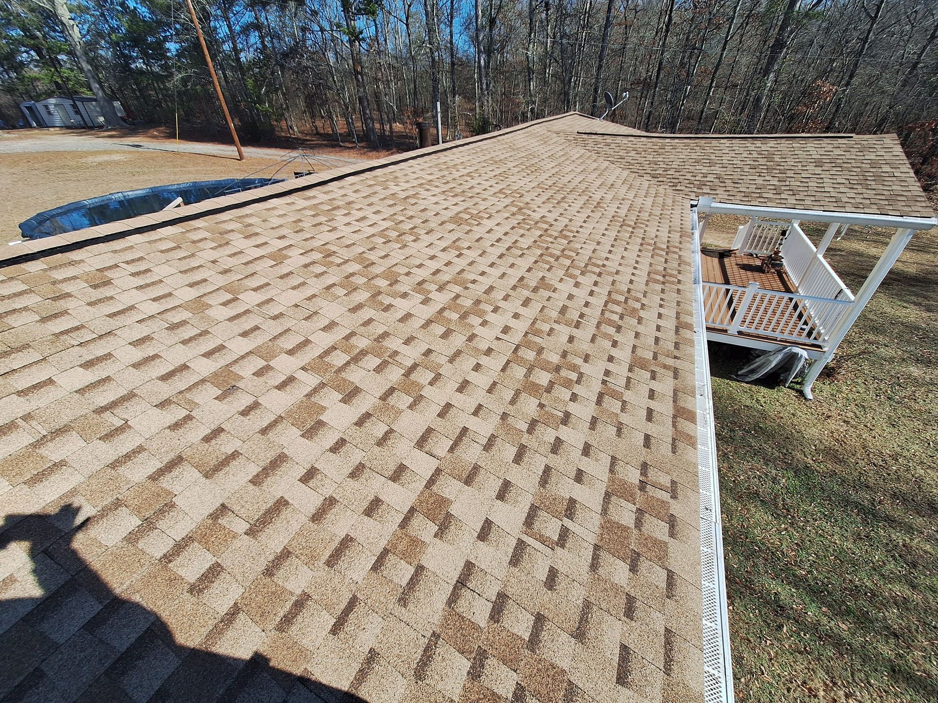Brown and tan shingle roof on a house with a small deck, trees in the background, and a pool.