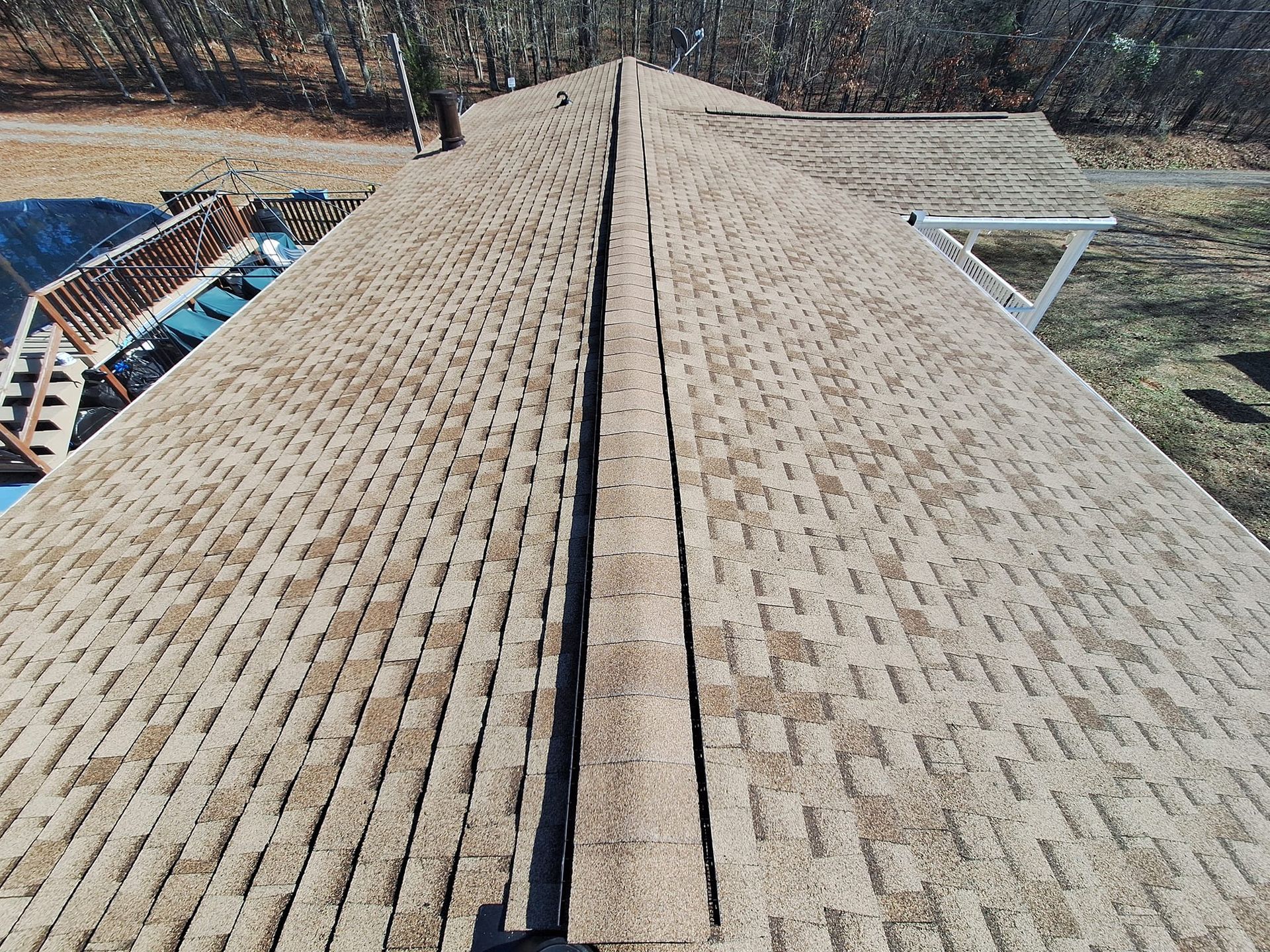 Aerial view of a brown shingled roof with a central ridge, surrounded by trees and a deck.