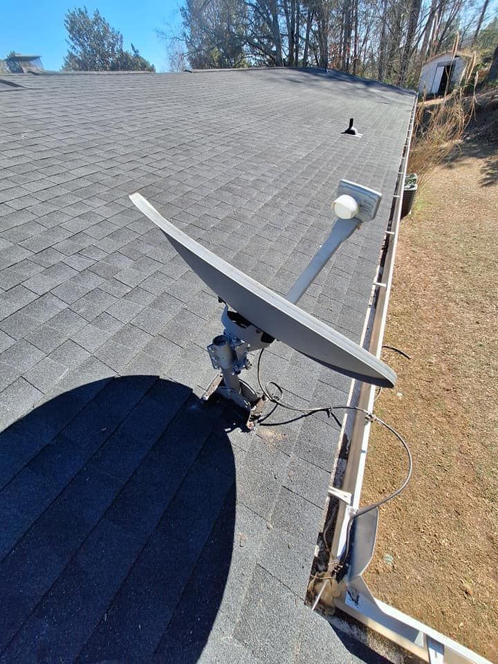 Satellite dish mounted on a dark shingled roof with a blue sky background.