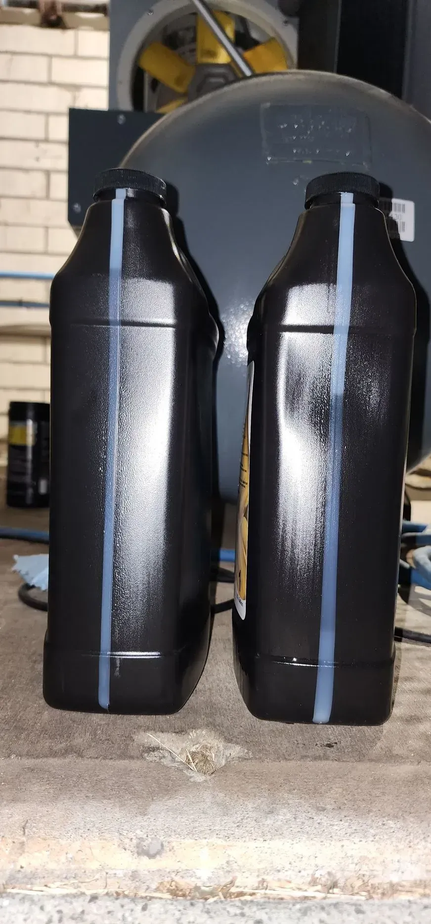 Two black oil containers side-by-side with grey stripes on a concrete floor near equipment.