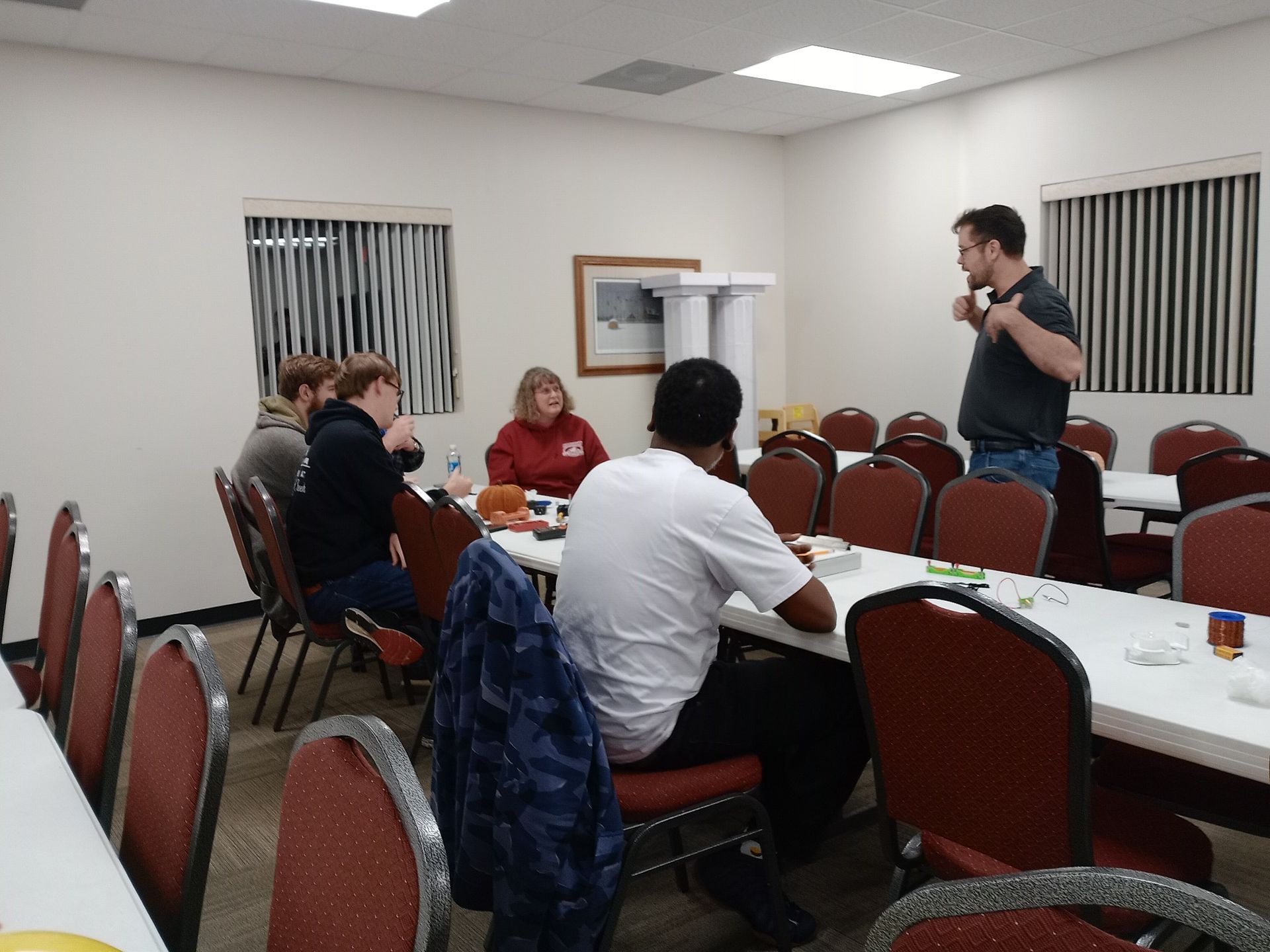 People at a table in a room. A man stands, talking. Others sit, some looking at a table with items.