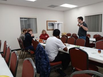 People at a table in a room. A man stands, talking. Others sit, some looking at a table with items.